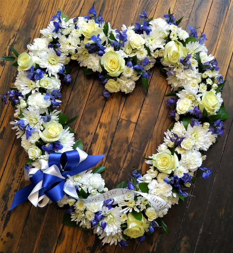 Heart-shaped wreath of white flowers with yellow roses, blue blooms, and a blue and white ribbon bow
