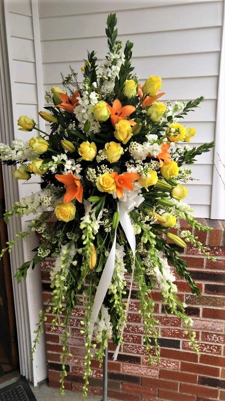 Tall standing spray with yellow roses, orange lilies, white flowers, and trailing green stems on an easel.