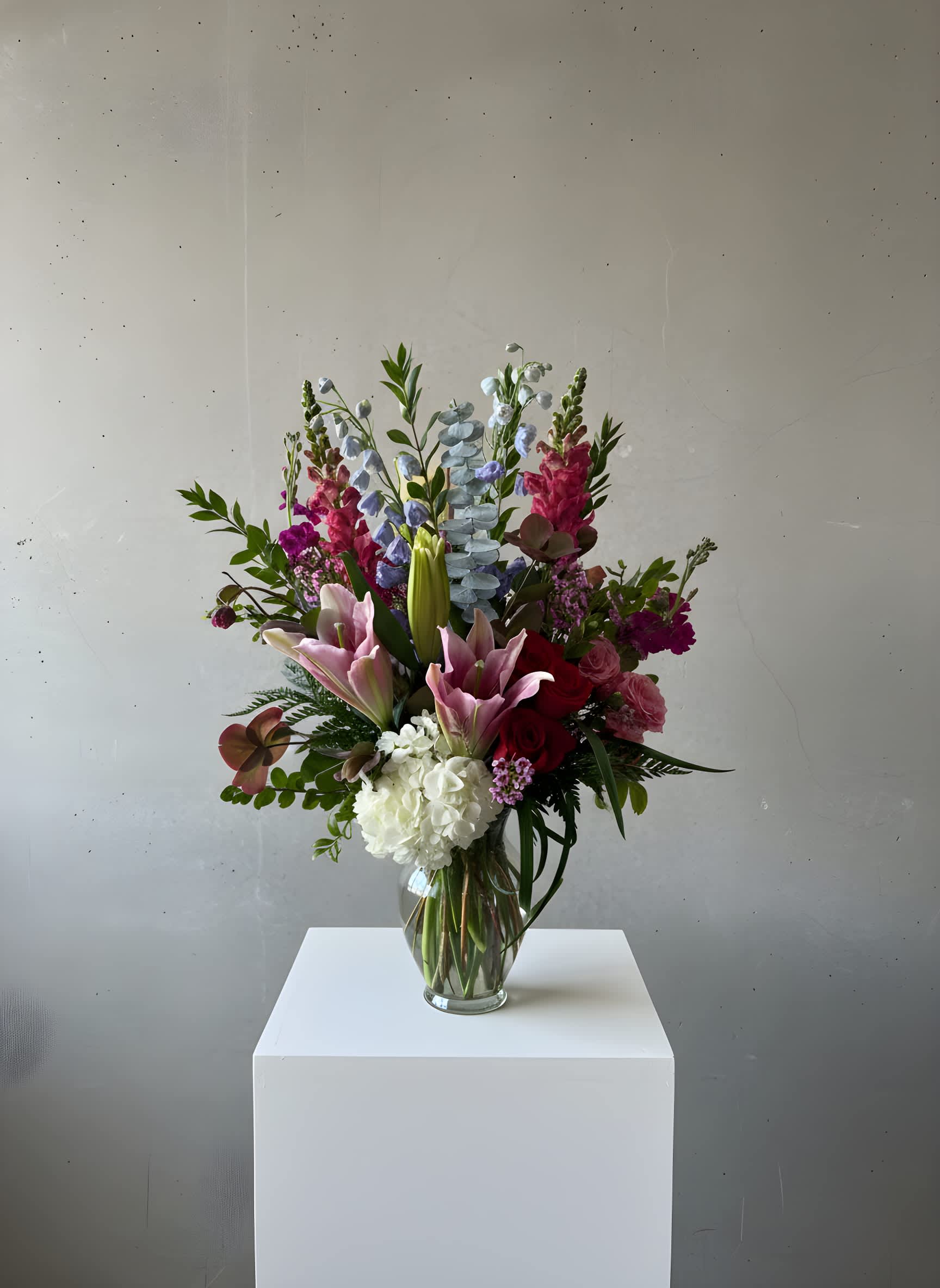 Tall mixed arrangement with pink lilies, red roses, white hydrangea, and blue delphinium in a clear glass vase.