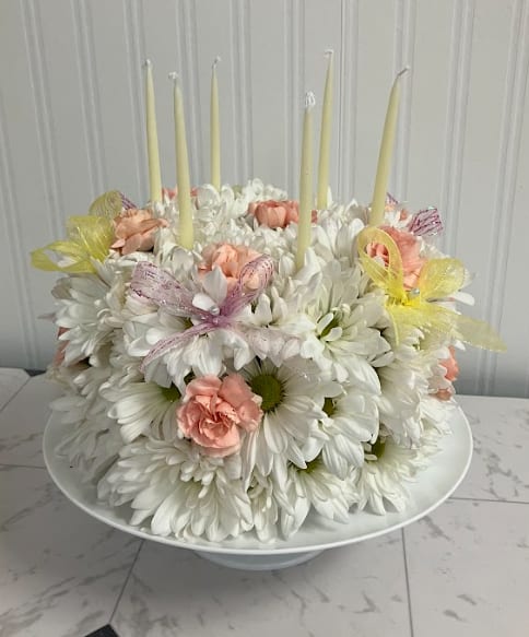 Let's Celebrate!  - Celebrate in bloom with our charming Floral Cake Arrangement, a sweet and whimsical design that looks almost good enough to eat. Crafted with fresh white daisies and soft peach mini carnations, this delightful creation is styled to resemble a frosted cake, complete with decorative pink and yellow ribbons and topped with candles for a festive touch. Perfect for birthdays, celebrations, or just because, this unique arrangement brings joy, color, and a creative twist to traditional floral gifting.