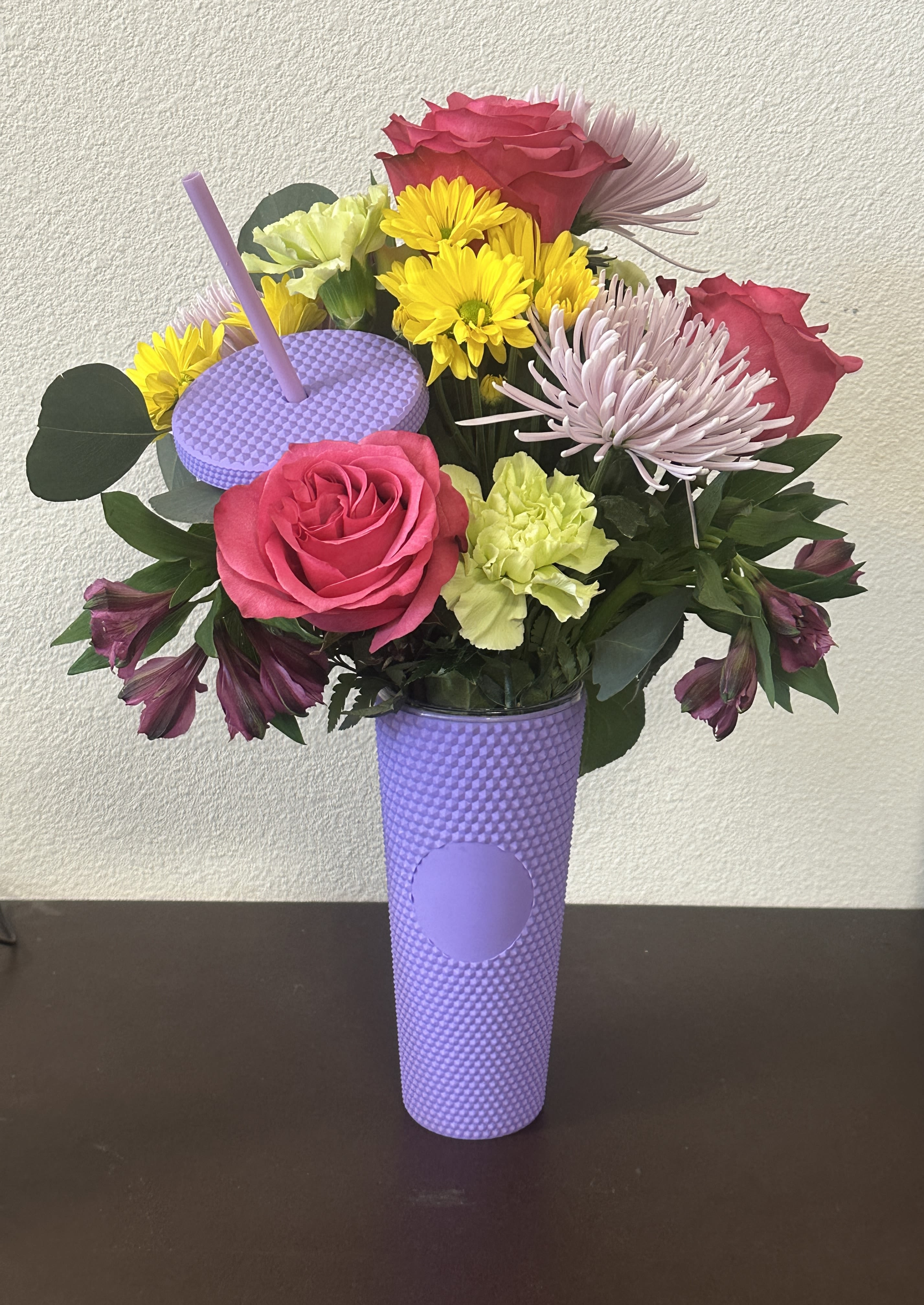 Bright mixed bouquet with pink roses and yellow daisies in a tall lavender tumbler cup with straw