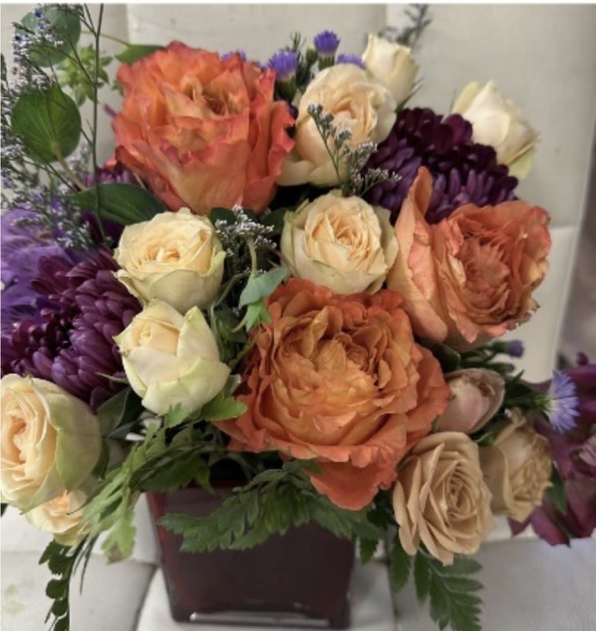 Arrangement of orange and cream roses with purple mums in a dark square vase
