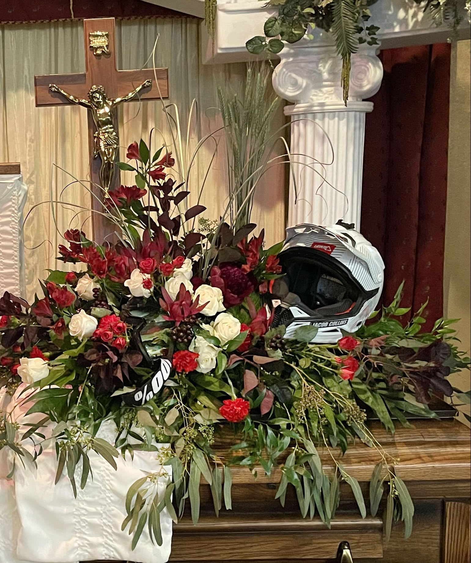 Red and white floral casket spray with roses and carnations displayed beside a white motocross helmet.