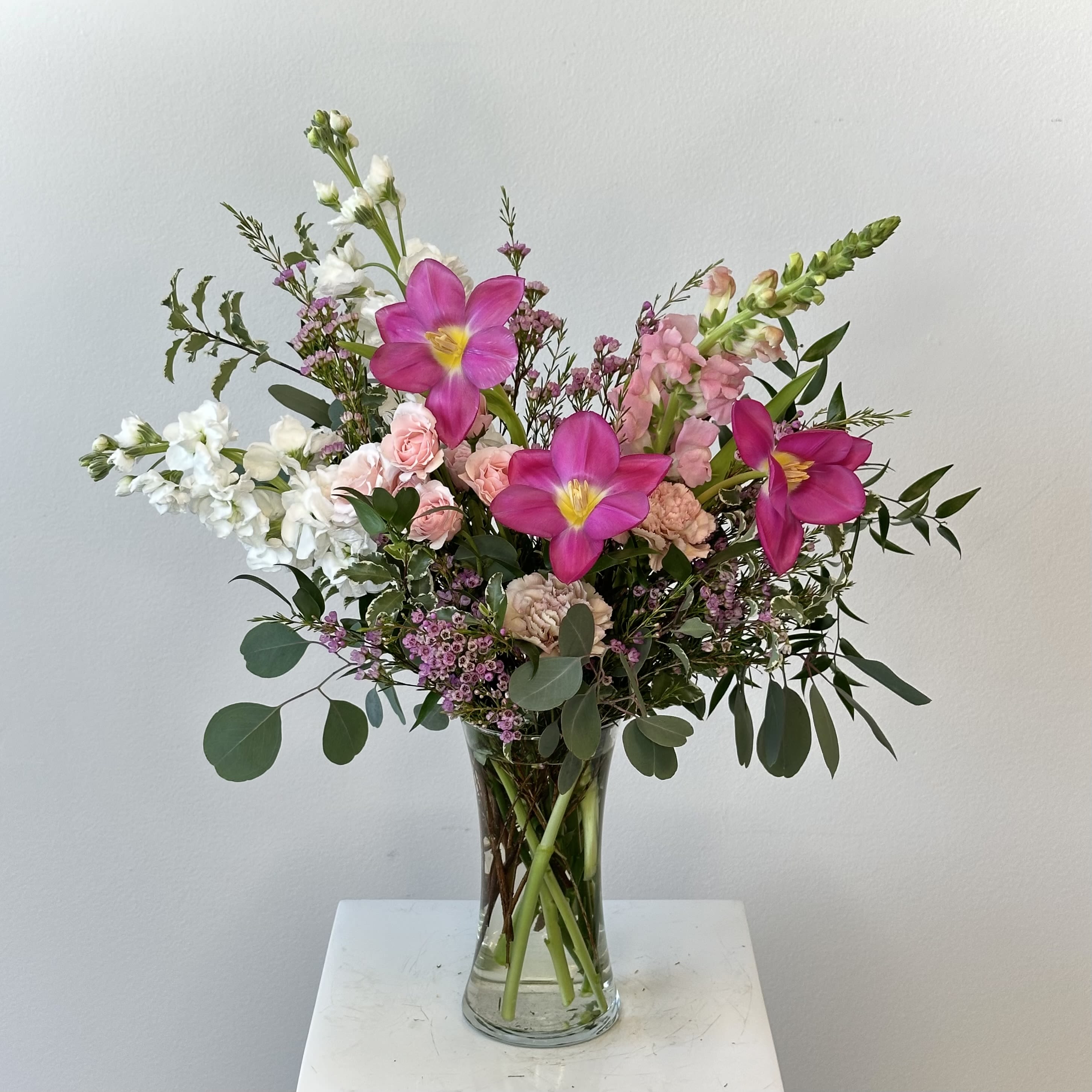 Wild Child - The Wild Child flower arrangement is a free-spirited and effortlessly beautiful design that captures the essence of natural, untamed elegance. Inspired by wildflower fields and organic garden style, this arrangement blends soft textures, airy movement, and fresh greenery to create a look that feels both relaxed and refined. Perfect for birthdays, celebrations, or everyday moments, Wild Child brings a breath of fresh air into any space.  Delicate tulips add a graceful, modern touch with their naturally curved stems and soft blooms, while fragrant white stock introduces height, fullness, and a subtle, sweet scent. Tiny wax flowers are thoughtfully woven throughout the arrangement, adding a light, whimsical texture that enhances its wild, meadow-inspired feel. Flowing eucalyptus provides a soft, silvery-green backdrop, creating movement and depth while complementing the gentle color palette.  Designed with an organic, loose style, the Wild Child arrangement feels effortlessly gathered yet artfully composed. Its balance of soft florals and fresh greenery makes it ideal for those who love a more natural, bohemian aesthetic. Whether given as a thoughtful gift or used as a centerpiece, this arrangement delivers understated beauty, texture, and charm, making it a timeless and versatile floral design.
