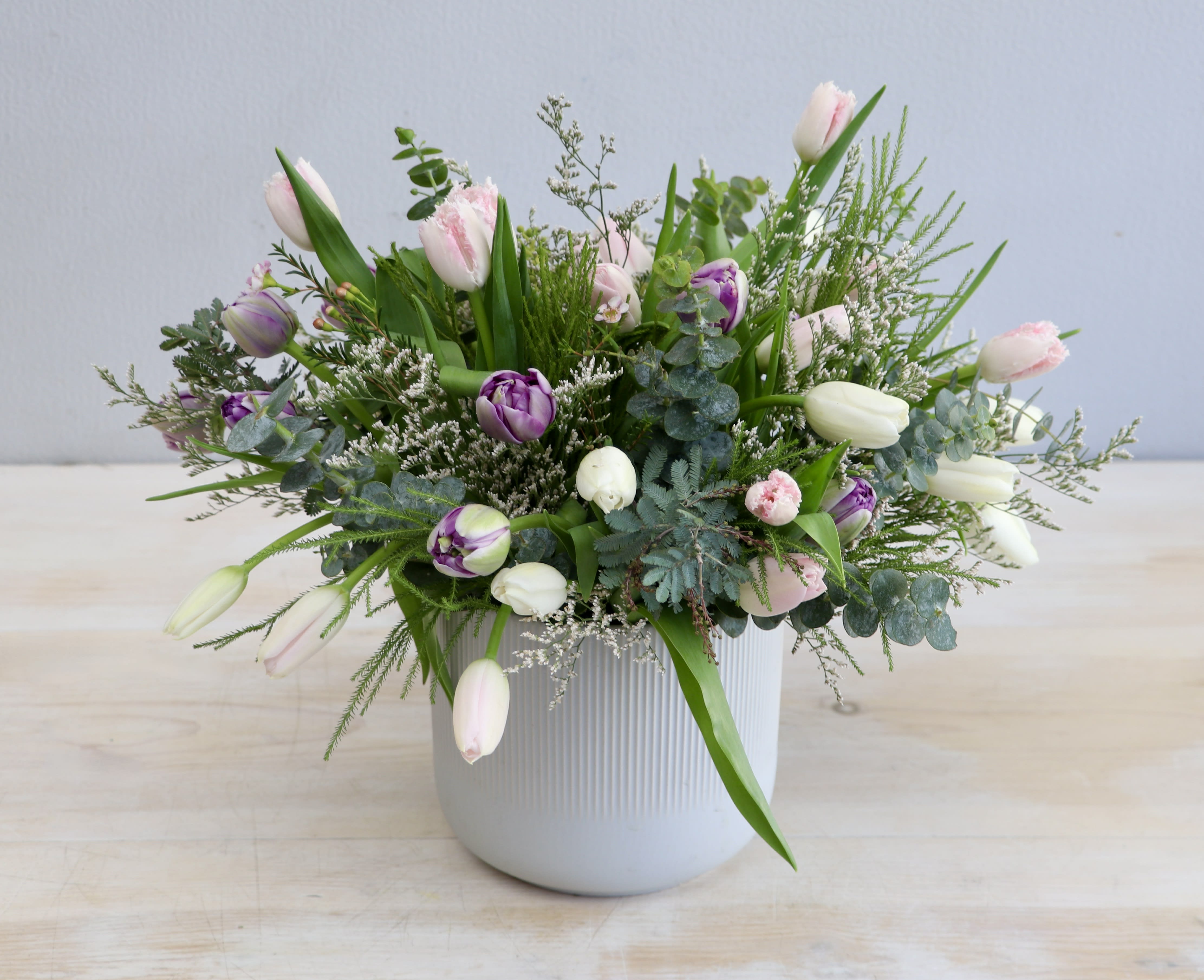 Low white ceramic pot with pink, purple, and white tulips arranged with airy greenery.