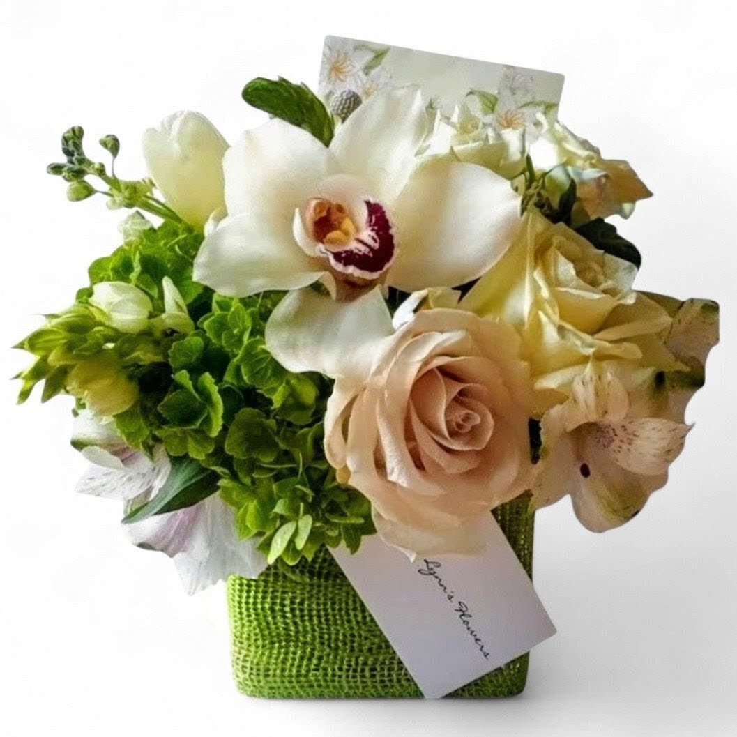 Small cube arrangement of white orchids, blush roses, and green blooms in a green vase with a card.