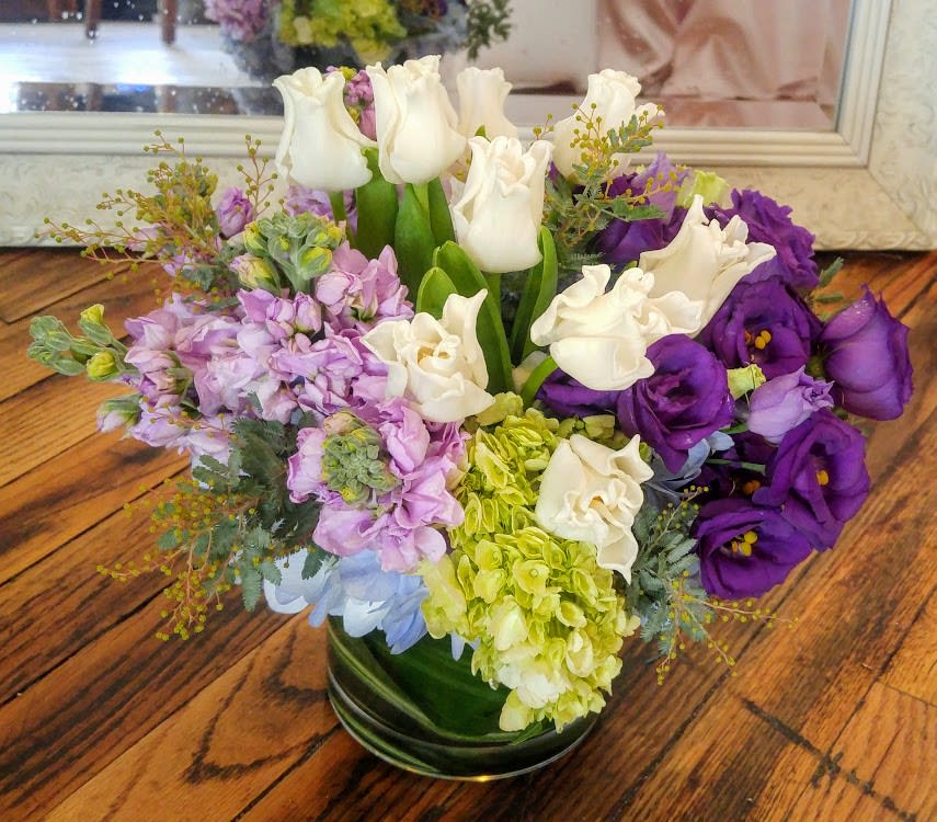 Low round arrangement of white tulips, green hydrangea, and purple lisianthus in a glass vase