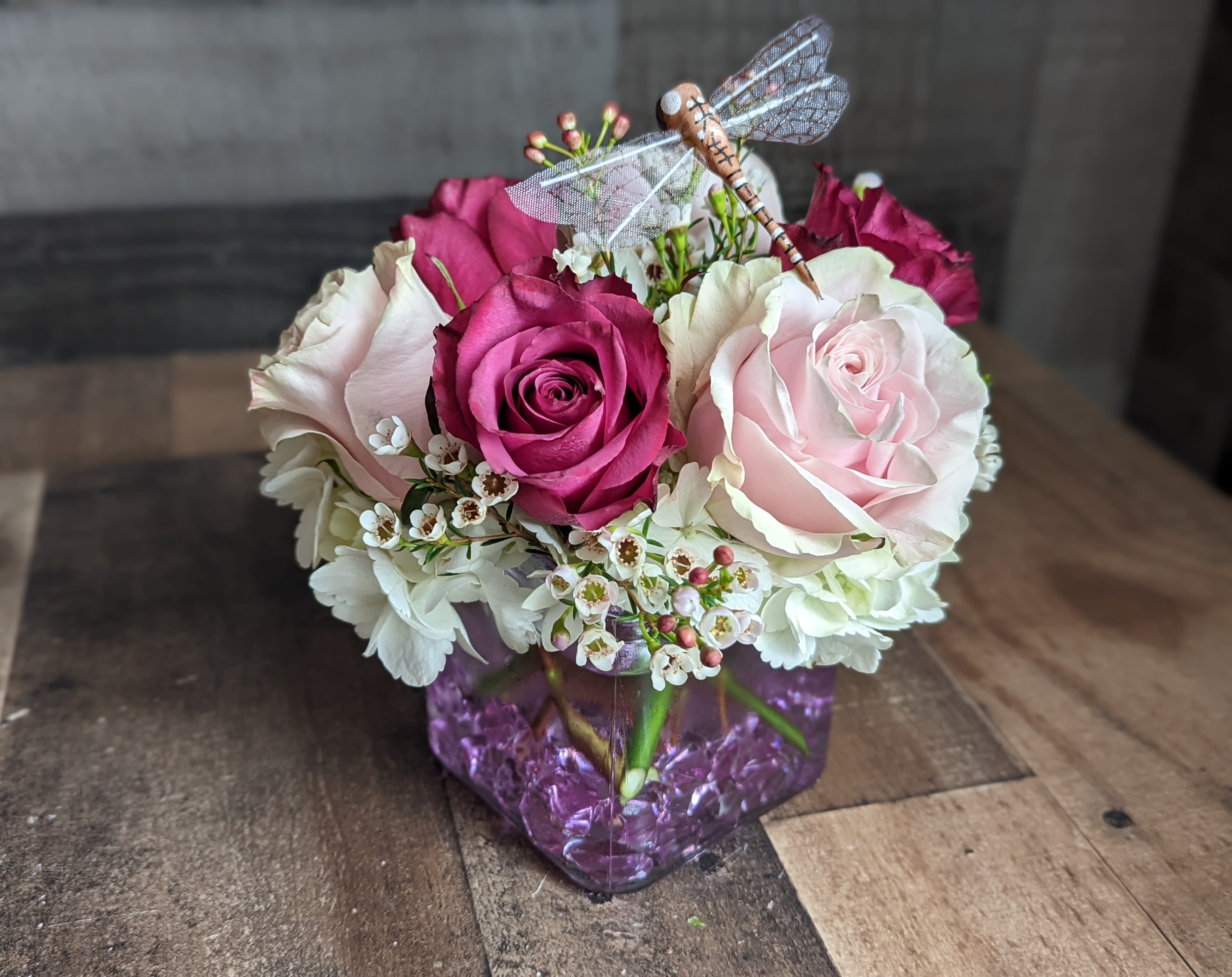 Short cube vase arrangement of pink and cream roses with white blooms and a decorative dragonfly pick.