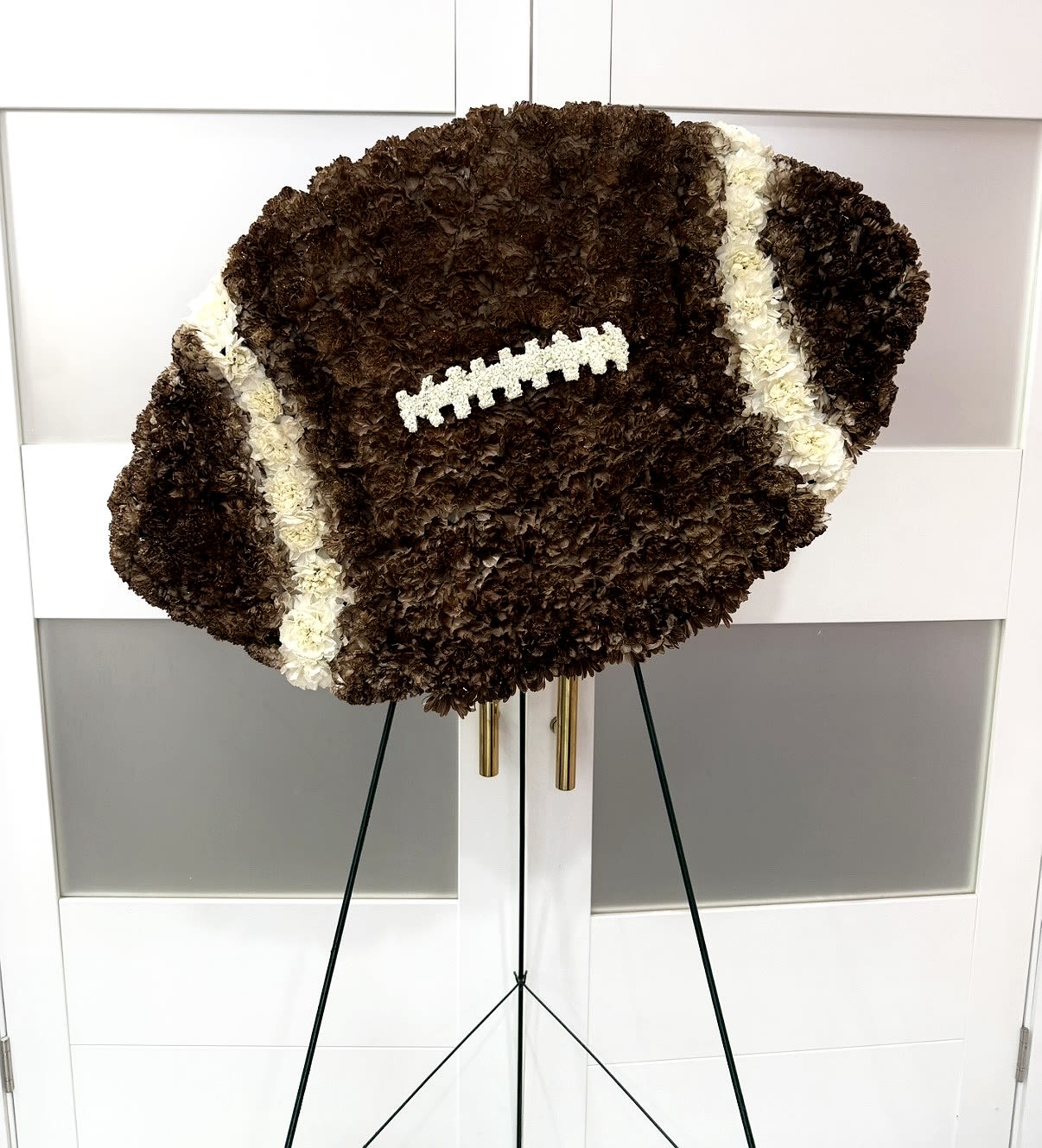Football- Sympathy Flower Tribute - Honor a life, passion, and love for the game with our custom-designed football floral tribute. Crafted with precision and care, this full-size football arrangement features rich, textured blooms in deep brown tones accented with crisp white detailing to resemble authentic laces and stripes.  Each piece is thoughtfully arranged to create a realistic and meaningful display—perfect for celebrating the memory of a sports enthusiast, athlete, or devoted fan. Set on an easel for a striking presentation, this tribute serves as a heartfelt symbol of dedication, teamwork, and lasting legacy.  Details:     •    Handcrafted with premium fresh flowers     •    Designed in the shape of a football with realistic detailing (approximately 24X36")      •    Displayed on a standing easel for services or memorials     •    Custom color options available upon request  A powerful and personal way to say goodbye while honoring a love that never fades.