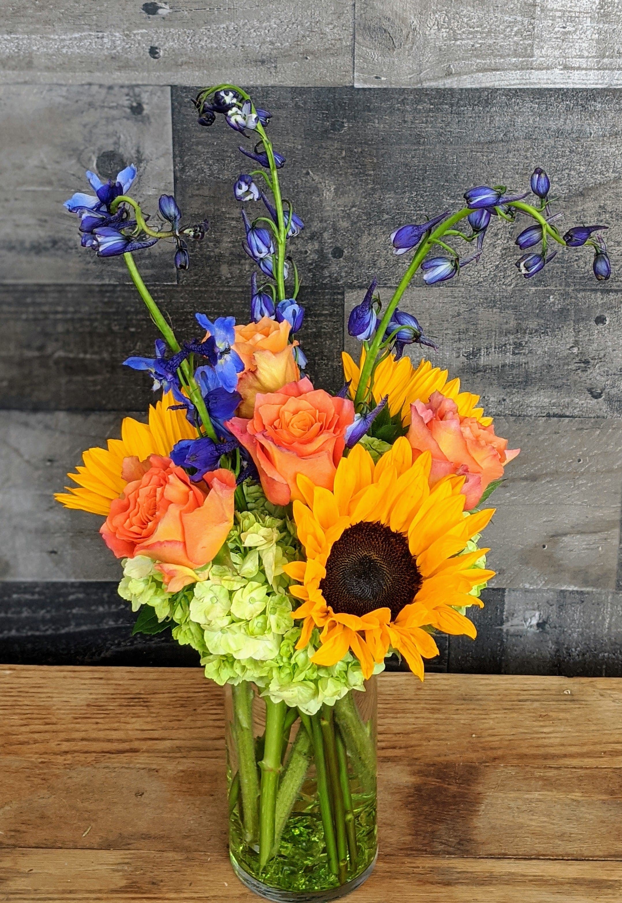 Tall glass vase with yellow sunflowers, orange roses, green hydrangeas, and blue delphinium.