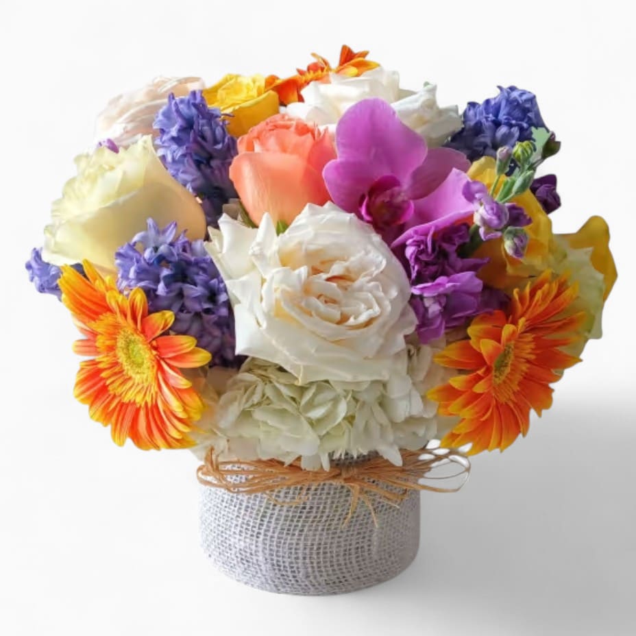 Low round arrangement of mixed white roses, orange gerbera daisies, and purple flowers in a wrapped glass vase