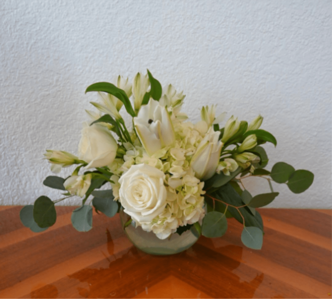 Low white arrangement of roses, lilies, and hydrangeas in a round glass vase