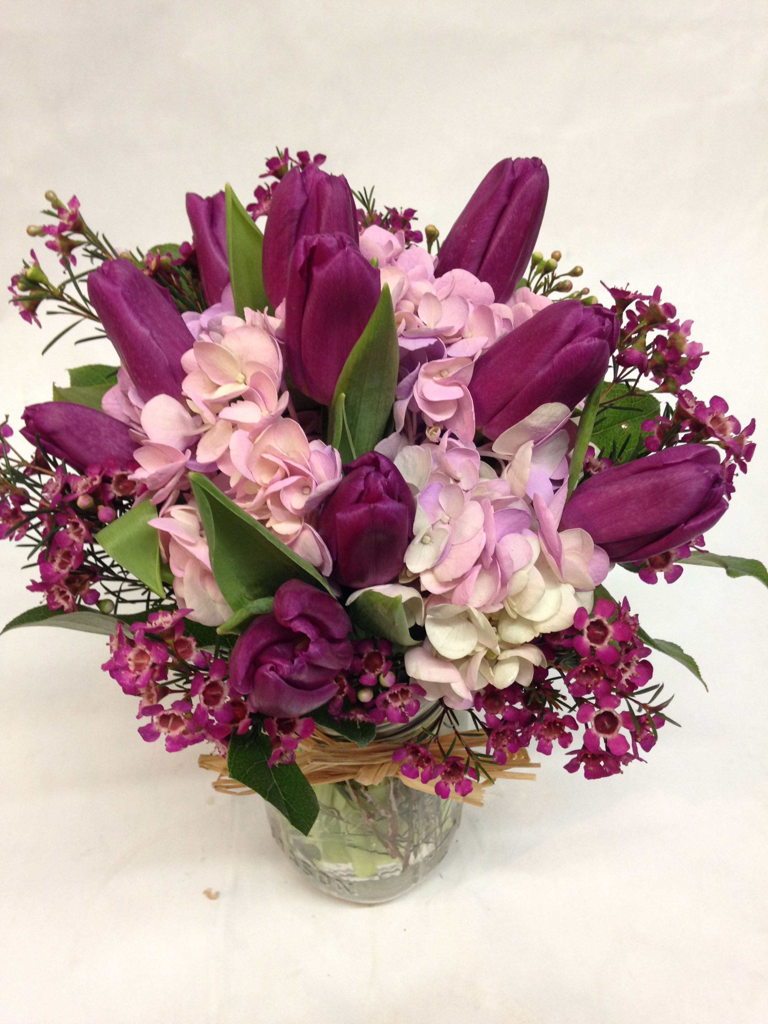 Purple tulips with pink hydrangeas and small blooms in a clear glass jar vase tied with raffia.
