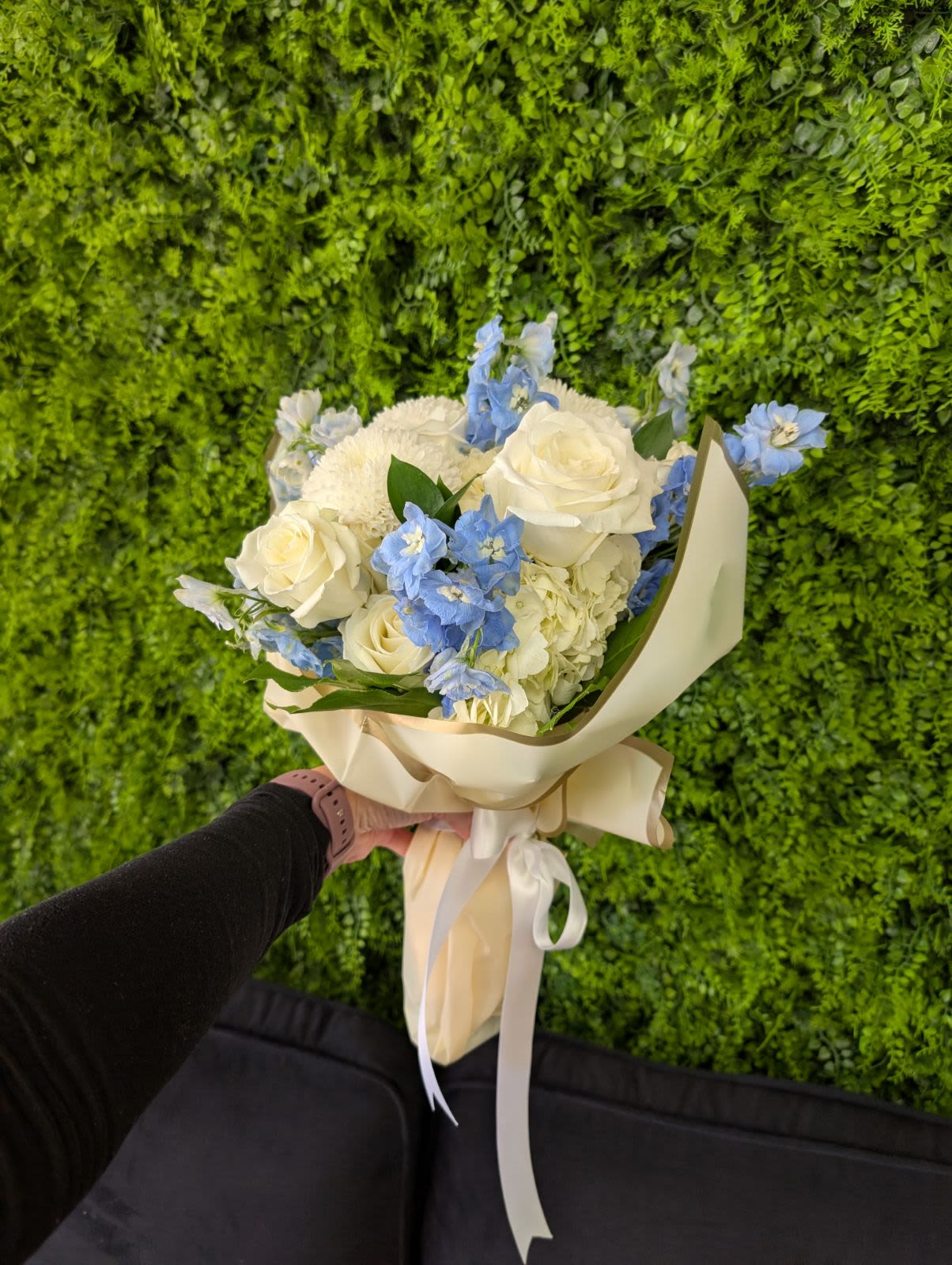 Hand-tied bouquet of white roses and blue flowers in cream wrap with ribbon, held against a green wall.