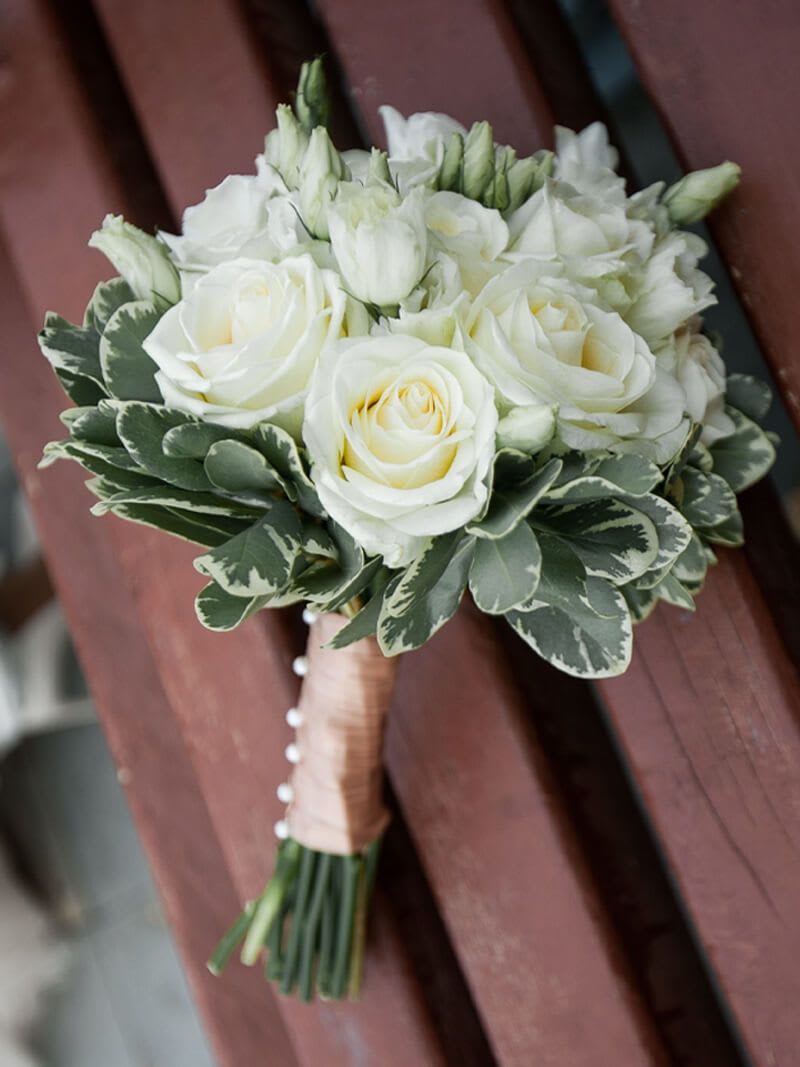 Elegant White Roses bouquet - A compact cluster of creamy white roses and delicate spray roses creates a soft, romantic focal point - the perfect bridal hand-tied bouquet for weddings and formal ceremonies. The neutral white-and-green palette with a whisper of peach reads timeless and refined, making it an ideal choice for the bride who wants classic elegance or for a mother-of-the-bride who prefers understated sophistication.  Creamy white roses and rose buds are tightly gathered into a classic round shape, framed by bold variegated foliage that forms a neat collar and adds crisp contrast and structure. The stems are bound with a peach-toned ribbon wrap and finished with a row of decorative pearl pins for a refined, bridal detail that shows up beautifully in photos. The compact size keeps the bouquet easy to hold throughout the service while still reading full and luxurious.  This bouquet is a graceful choice for traditional ceremonies, formal receptions, or as a complimentary bouquet for bridesmaids seeking a coordinated look. Hand-tied for a comfortable grip and lasting structure, it arrives ready to carry - lightly mist stems with water before the event and keep in a cool place until the processional to maintain peak freshness.