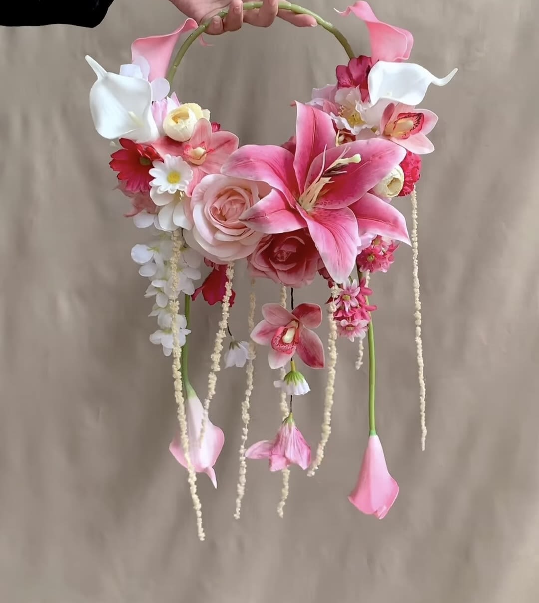 Bridal Flower Hoop Bouquet - STANDARD: Silk Faux Flowers | DELUXE: Fresh Flowers - STANDARD: SILK FAUX Flowers $190 DELUXE: FRESH Flowers $245  A circular hoop bouquet that reads like a wearable sculpture-this Bridal Flower Hoop brings romantic, modern flair to wedding florals. Perfect as flowers for weddings, the design places a lush band of pink lilies, blush roses, peach calla lilies, orchids, and delicate daisy-like blooms along the lower half of a hoop bouquet frame, creating a soft, feminine focal point that complements bridal and bridesmaid looks alike.  The palette of pink, white, and peach is balanced so the creamy whites lift the composition while the pinks and peaches deliver warmth and depth. Long trailing elements and a few hanging blossoms add gentle movement and a cascading silhouette that photographs beautifully down the aisle and in portraits. The hoop bouquet frame is hand-held for easy carrying during processions and offers a striking, contemporary alternative to traditional posy or cascade bouquets.  Made to make the recipient feel seen and celebrated, this hoop suits brides seeking a whimsical yet modern statement or attendants who want a coordinated, picture-ready accessory. Its compact, wearable form keeps it comfortable to hold but visually impactful for close-up photos; if you have specific color accents or placement needs for your ceremony, we can accommodate those requests when you order.