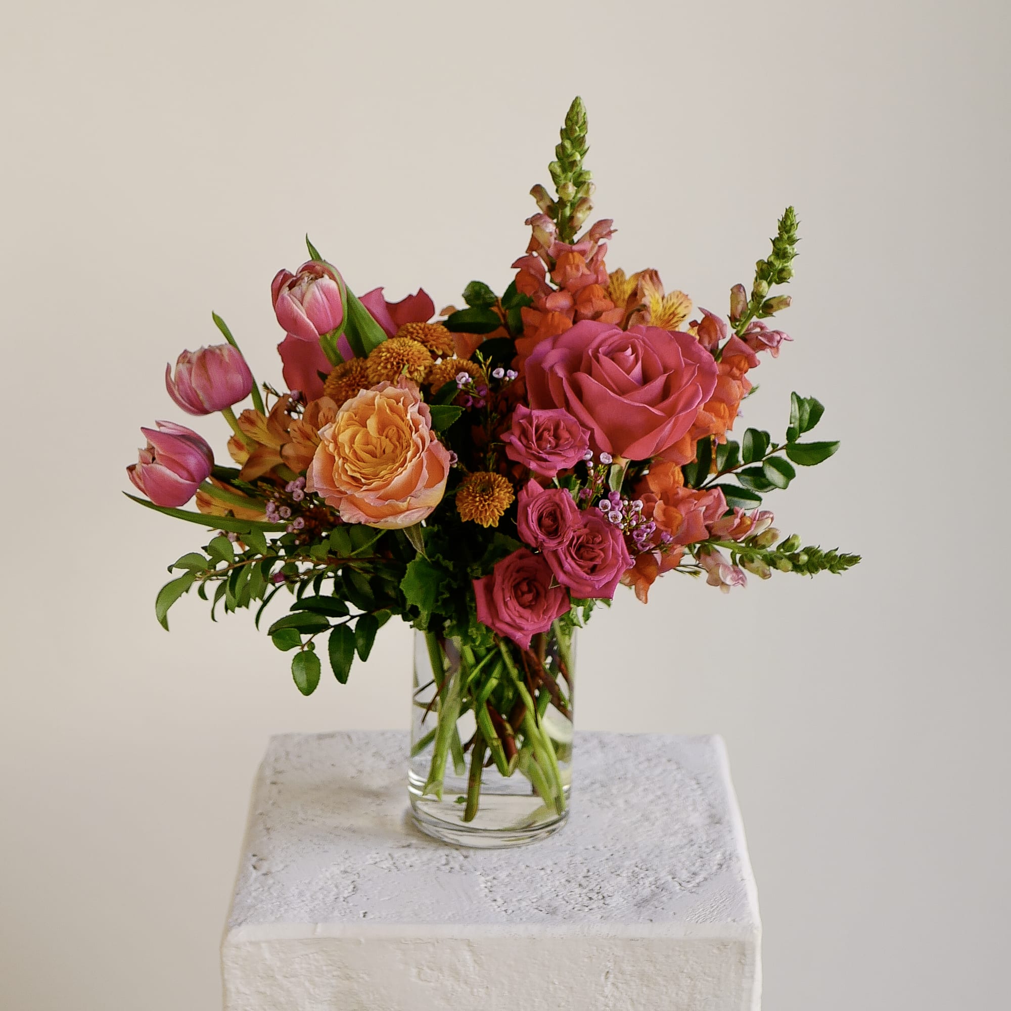 Gem Star - Gem Star is for when you want flowers that actually stop people in their tracks. Hot pink roses, vibrant orange roses, coral tulips, snapdragons, alstroemeria, and mums — all arranged in a loose, garden-style design with waxflower and fresh greens woven throughout. Presented in a 4x6" clear glass cylinder vase. Arrangement will be delivered approximately as pictured, about 14" tall and wide with flowers.  Perfect for birthdays, celebrations, or anyone who loves bold color done beautifully.