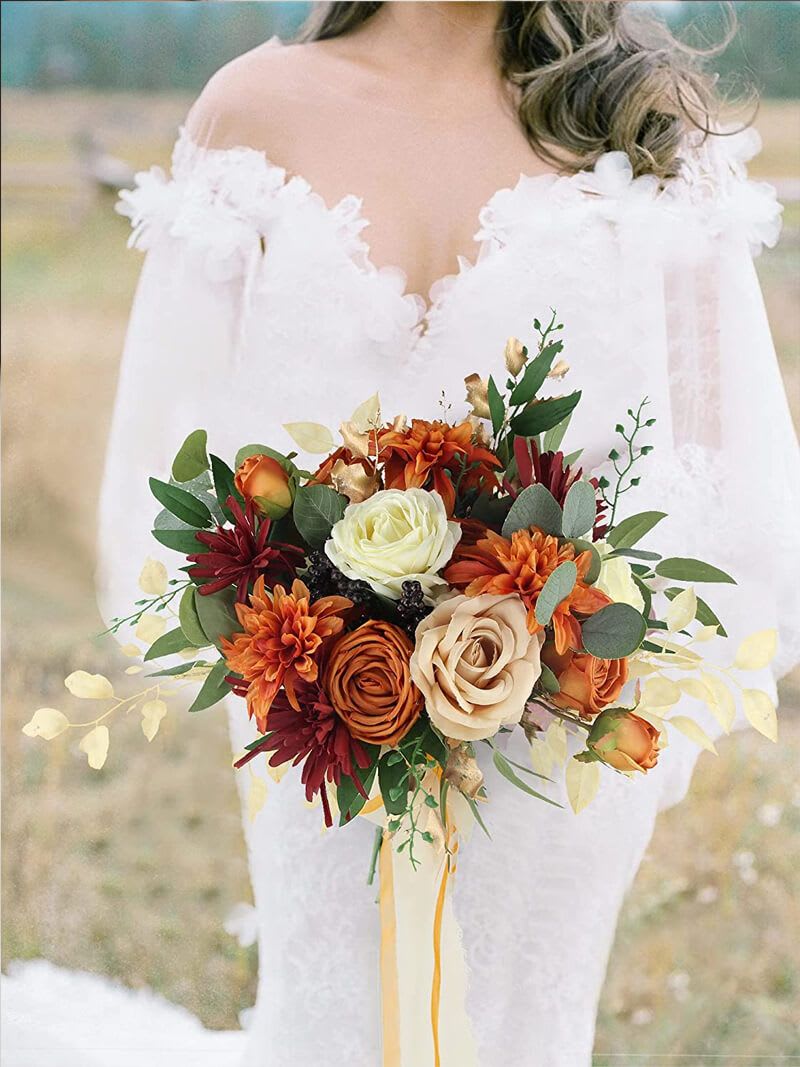 Mixed Flower Bouquet - A warm, hand-held wedding bouquet that feels like autumn sunlight-perfect for brides seeking a romantic, slightly rustic look for their ceremony. This mixed flower bouquet for weddings blends mixed color roses and chrysanthemums in orange, red, white and peach tones to create a rich, rounded silhouette that reads beautifully in photos and next to a lace gown.  Large tan and ivory roses sit at the heart of the design, surrounded by rust orange and deep burgundy chrysanthemum-style blooms and varied foliage for texture and movement. Pale, delicate accents extend just beyond the rounded shape to keep the arrangement feeling natural and effortless. Long satin ribbons in soft yellow and ivory trail from the base, adding a flowing, timeless finish.  Made to complement fall and vintage-inspired weddings, this bouquet is a meaningful way to make the person you're giving it to feel truly seen on their day. Sized as a classic bridal bouquet and hand-tied for secure handling, care is simple-keep stems in water until just before the ceremony and mist lightly to maintain freshness.