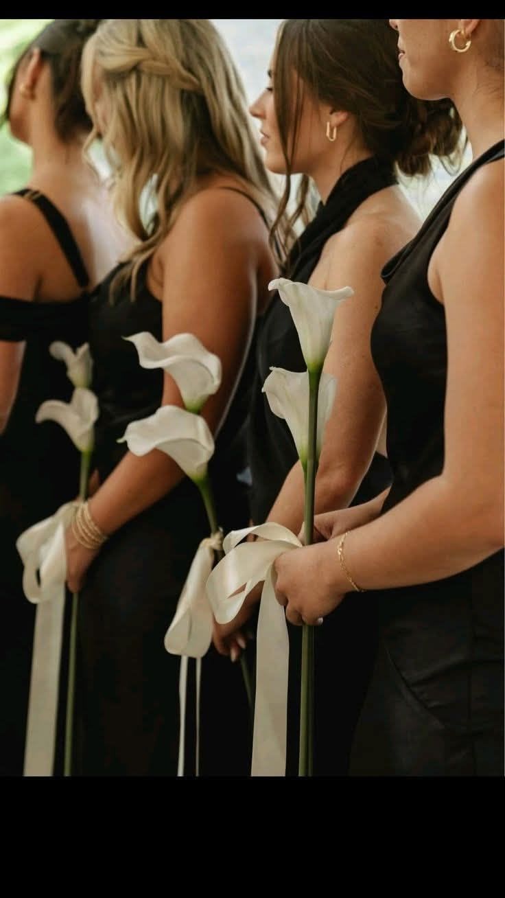 Calla Lilies Bridesmaid Bouquet - White | STANDARD: Silk Faux Flowers | DELUXE: Fresh Flowers - STANDARD: Silk Faux Flowers $40 DELUXE: Fresh Flowers $45 A line of graceful white calla lilies makes a quietly modern statement for bridesmaid bouquets, wedding parties, or refined sympathy settings. These long-stem calla lilies carry a sleek, ceremonial feel-their natural curves and bright white spathes stand out against darker dresses and lend a clean, contemporary note to any formal occasion.  Each bouquet is a single long-stem calla lily tied near the base with a trailing white ribbon, letting the smooth green stem and elegant bloom remain the focus. The simple black, white and green palette keeps the look cohesive with tuxedos, black bridesmaid gowns, or monochrome wedding themes, while the minimal hardware ensures a comfortable, easy-to-carry design.  Perfect for brides seeking a minimalist, sophisticated look or for giving as a tasteful sympathy spray alternative, these stems arrive ready to carry and photograph.   Handle gently by the stem, keep blooms in water until the moment they're needed, and order in the quantity that matches your party for a coordinated ceremony presentation.