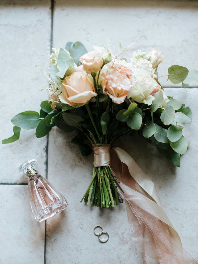 Made For You - A hand-tied wedding bouquet that reads like a quiet love letter-peach and ivory roses surrounded by soft, smaller blooms and rounded eucalyptus make this bridal bouquet perfect for weddings and romantic celebrations. Made for the person you're marrying, it brings a timeless, softly styled look that complements classic ceremonies and intimate receptions.  The stems are tightly wrapped in blush and ivory satin ribbon, finished with long, trailing sheer ribbons for elegant movement as she walks down the aisle. The palette of white, peach, pink and green creates a muted, pastel harmony; the mix of roses and delicate filler flowers gives both lush volume and gentle texture. Styled here with a faceted glass perfume bottle and simple wedding bands to evoke the bridal moment.  Ideal for brides seeking a romantic, classic bouquet that photographs beautifully and feels effortless to carry. Measures as a hand-tied cluster sized for ceremony use; care for roses by keeping stems hydrated until delivery and storing in a cool, shaded spot before the vows.