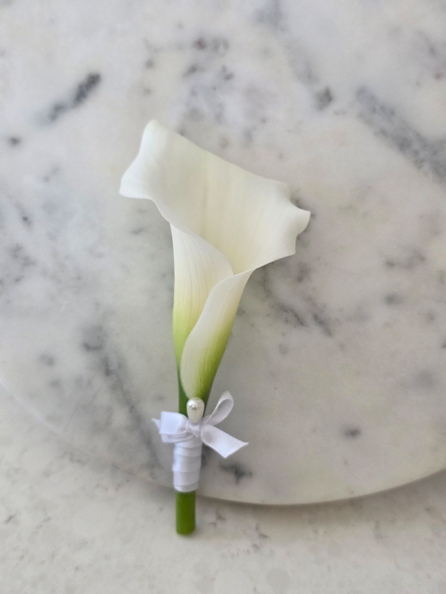 Calla Lily Boutonniere- White - A single soft-white calla lily boutonniere with a whisper of green at the bloom's base offers a refined, modern accent for formal occasions. Ideal for prom, weddings, and retirement celebrations-or as a respectful touch for sympathy and funeral services-this boutonniere reads clean and intentional so the person wearing it will feel quietly honored and seen.  The slender stem is neatly wrapped in smooth white satin ribbon and finished with a small bow, then secured with a decorative pearl pin for a polished, timeless look. Its minimalist silhouette and neutral palette pair beautifully with suits or gowns without competing with other floral choices, making it an elegant choice for wedding parties, graduation events, or any moment that calls for understated sophistication.