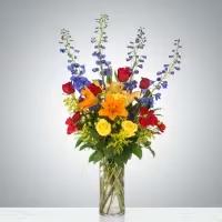 Vibrant Harmony Vase - A bold and modern vase arrangement bursting with color and texture. This striking design features rich red roses and red spray roses, bright orange lilies, cheerful yellow roses and solidago, and vibrant blue delphinium. The combination of warm and cool tones, accented with lush greens, creates a lively, contemporary display perfect for celebrating any joyful occasion.