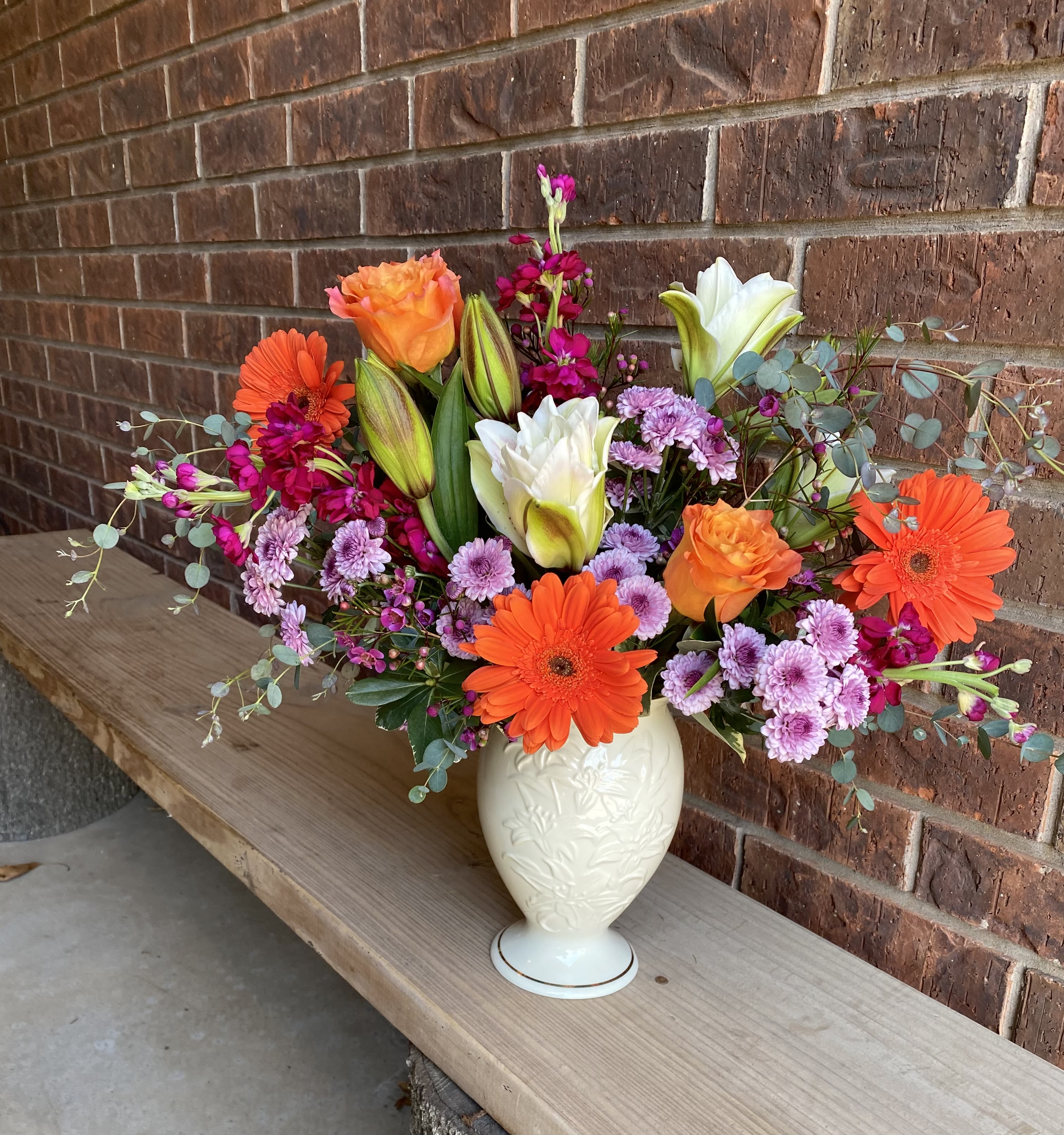 Sunrise - A burst of sunrise hues-vibrant orange gerbera daisies and roses paired with crisp white lilies-makes this flower arrangement a joyful choice for a birthday bouquet or a cheerful "thinking of you" gift. The lively composition also suits anniversaries or congratulations, giving the person you're thinking of an immediate lift with bright, saturated color and a welcoming, celebratory mood.  Soft lavender chrysanthemums and clusters of hot pink stock weave through the arrangement, while lily buds add graceful vertical texture and promise of evolving blooms. All flowers are arranged in a cream ceramic vase with an embossed floral pattern, a sturdy, tactile base that complements the warm tones and will sit beautifully on a kitchen table or office desk.  Send this when you want someone to feel genuinely seen and celebrated-it's the kind of bouquet that reads as thoughtful and exuberant at the same time. The vase arrives ready to display; trim stems and refresh the water every 2-3 days to keep blooms looking their best.