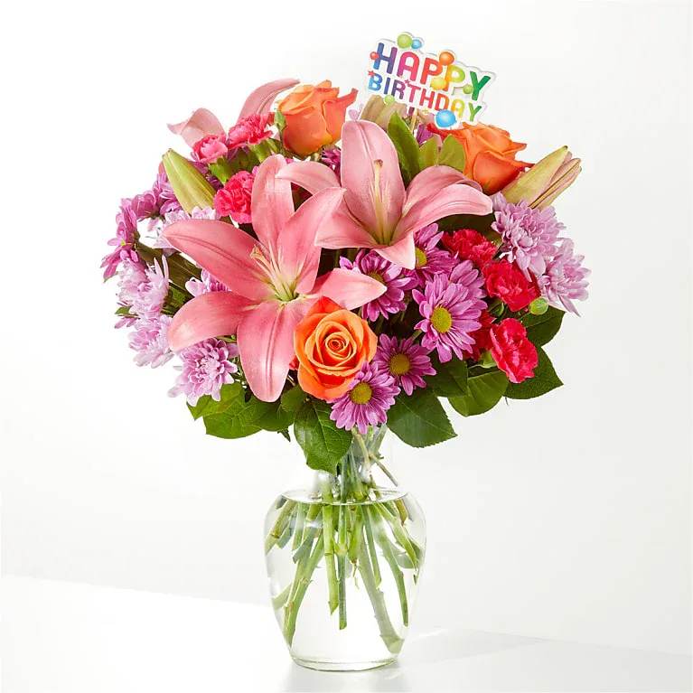Mixed bouquet of pink lilies, orange roses, and purple mums in a clear glass vase with a Happy Birthday pick.