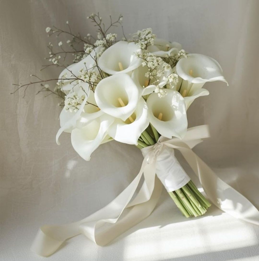Calla Lilies Bridal Bouquet - White - A cluster of pure white calla lilies arranged in a tight, hand-tied formation creates an unforgettable bridal bouquet that reads as both modern and timeless-ideal for a wedding or an elegant anniversary. The clean silhouettes of the callas give a sleek, architectural feel while the small white filler flowers woven through the arrangement add a soft, cloudlike texture that keeps the look romantic rather than austere. The overall impression is calm, sophisticated, and unmistakably ceremonial-the kind of bouquet that makes the person you're honoring feel beautifully seen.  Each stem is neatly wrapped in white fabric and finished with a long ivory satin ribbon that trails gracefully, perfect for carrying down the aisle or photographing in soft natural light. Color is limited to white and green for a classic palette; materials include calla lilies, delicate white filler blooms, a white fabric stem wrap, and an ivory satin ribbon. This compact, hand-tied size is easy to hold throughout the ceremony and will remain fresh with routine care-keep stems in water until the moment of use and store in a cool, shaded spot before presenting.