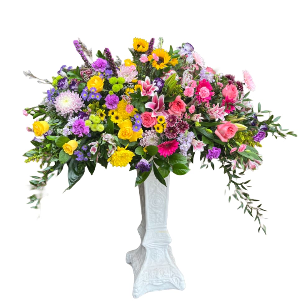 Spring Garden Tribute - A wide, overflowing display of spring color-perfect as a sympathy or memorial arrangement or as a grand ceremony piece. This spring garden tribute is a large, elongated flower arrangement designed for a white pedestal stand, combining pink and yellow roses with hot pink gerbera daisies and bright sunflowers to create an immediate, uplifting impression. Lavender and purple accents cool the palette, giving the person you're honoring a balance of warmth and calm that feels both celebratory and dignified.  The design mixes stargazer-style lilies, varied chrysanthemums and button-style blooms, snapdragons, alstroemeria, asters and other small florals for texture and movement; some stems arch outward to create a sweeping, garden-like silhouette. The overall shape is lush and horizontal, intended as a statement piece on an ornate white pedestal stand. Sized for large events-weddings, memorial services, or any occasion calling for a formal tribute-this arrangement delivers vivid color and layered texture that will make the recipient (or everyone who sees it) feel genuinely seen and remembered.
