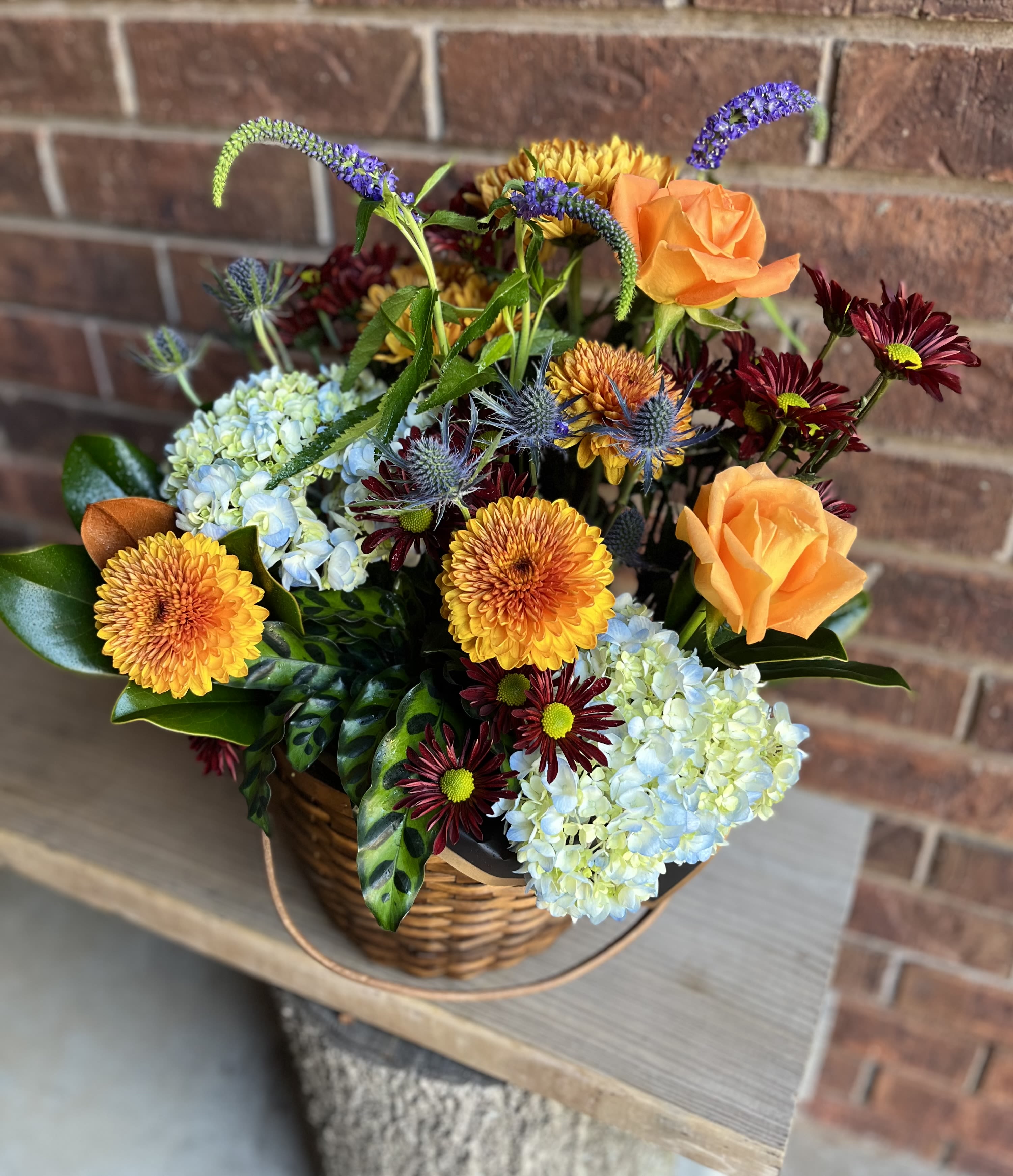 Autumn Sun - A rustic, autumnal centerpiece arranged in a low woven basket, this seasonal bouquet of plants and fresh florals makes a cozy gift for birthdays, housewarmings, or to lift someone's spirits. The mix of orange roses and golden chrysanthemums creates a warm, welcoming glow, while pale blue hydrangeas and deep burgundy accents give the design depth-perfect as a birthday bouquet or a thoughtful "thinking of you" arrangement.  Textural details-spiky blue thistle-like blooms and slender purple flower spikes-lend a slightly wild, garden-inspired feel that still looks tidy on a mantel or dining table. The arrangement pairs warm oranges and yellows with cool blues and purples for contrast, finished in a sturdy woven basket that arrives ready to display.  This is the kind of gift that makes the recipient feel genuinely seen: cozy, seasonal, and carefully composed. The low profile fits easily into home settings; water and care are standard for mixed-cut flowers-keep in a cool spot, refresh the water every other day, and remove wilted stems to prolong enjoyment.