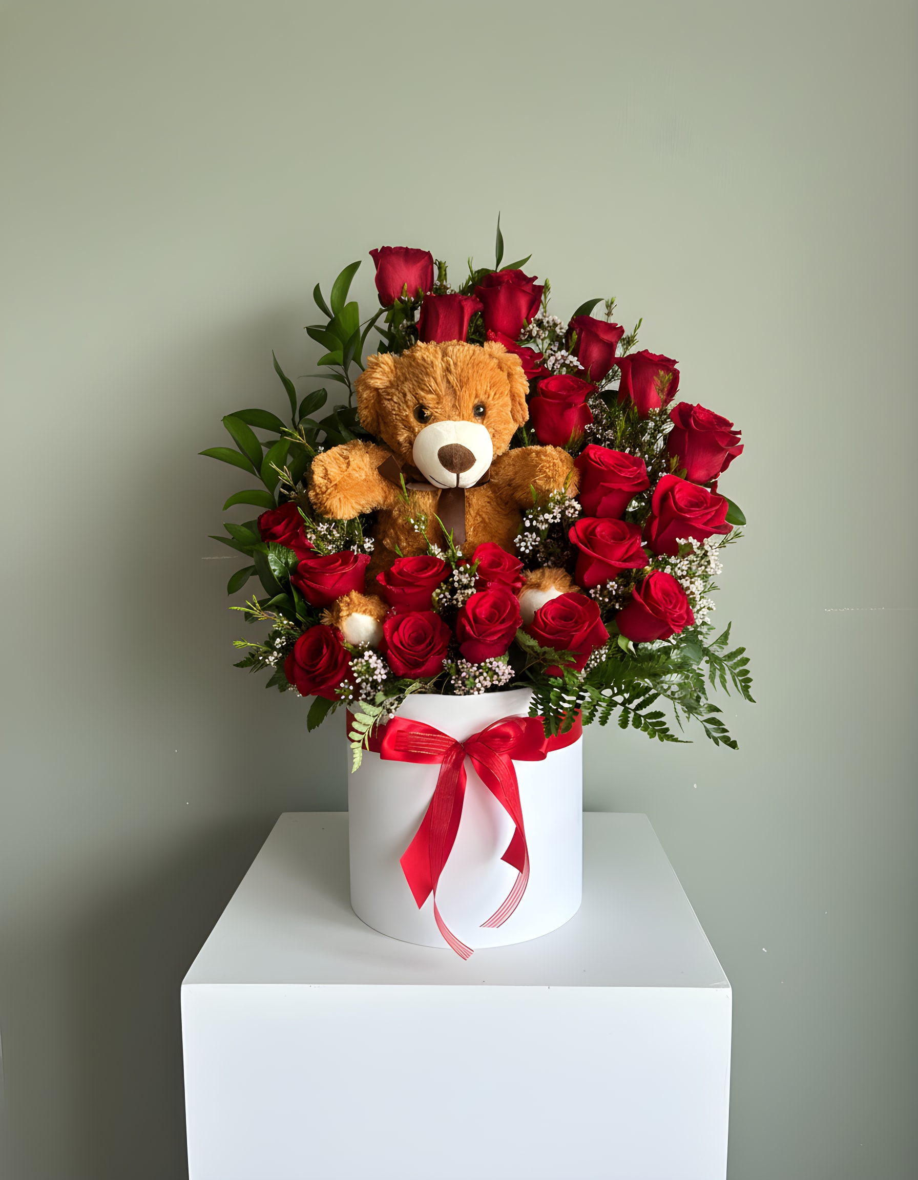 Round box of red roses with small white blooms and a teddy bear nestled in the center.