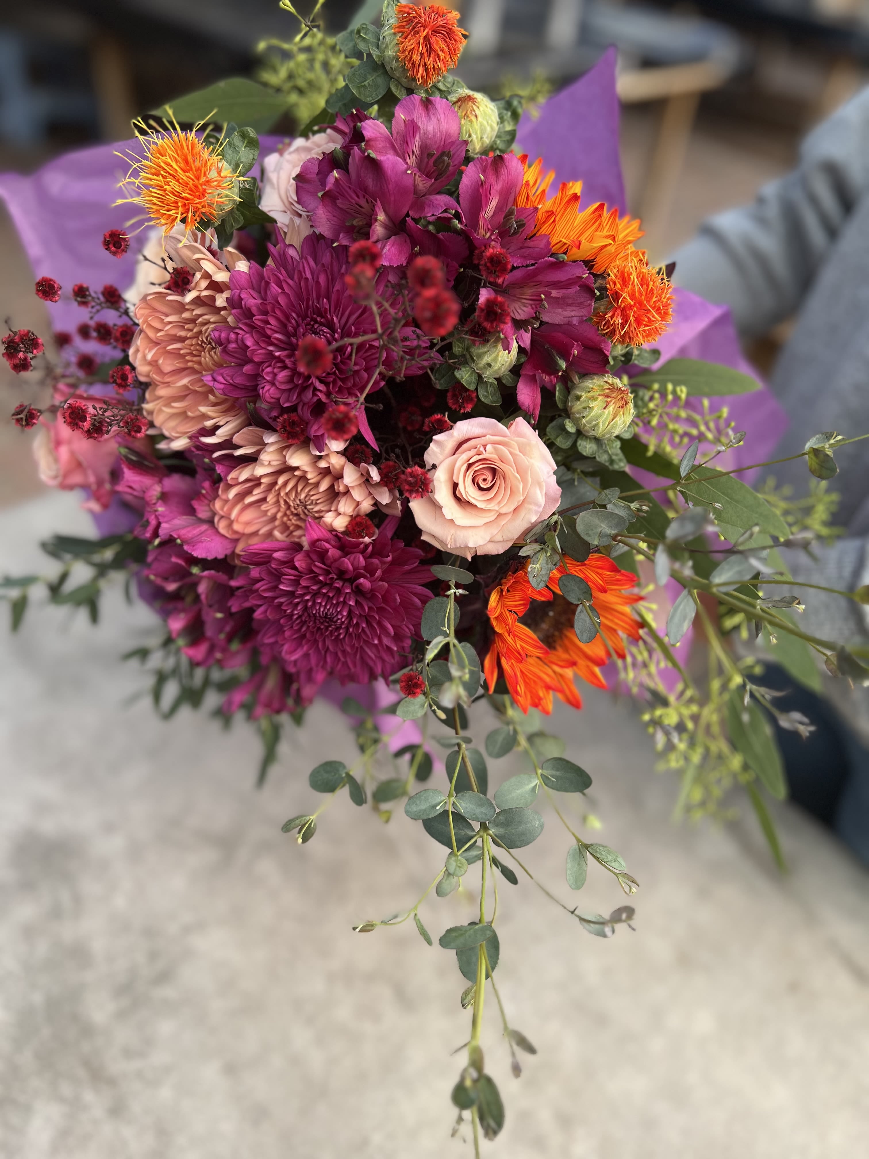 Hand-tied bouquet of purple mums, pink roses, and bright orange blooms with trailing greenery.