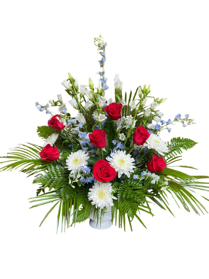 Patriotic Urn Pot - A fan-shaped tribute of red roses, white chrysanthemums, and soft blue delphinium creates a dignified sympathy or patriotic urn pot that reads both reverent and celebratory. This flower arrangement for memorials or patriotic observances places the occasion front and center: the person you're honoring will be remembered with classic red, white, and blue blooms arranged in an altar-style silhouette that speaks to respect and remembrance.  Clusters of velvety red roses form strong focal points across the center and sides, balanced by cushion-style white chrysanthemums and vertical spikes of light blue delphinium that lift the composition upward. Broad, fanning greenery frames the arrangement and gives a structured, wide base; the flowers are arranged in a simple white vase that keeps the focus on color and form. The overall proportions favor a tall, formal presence suitable for display on an urn table or memorial pedestal.  Designed to convey solemnity and patriotic sentiment, this urn pot is a meaningful way to honor loved ones at funerals, remembrance services, or national observances. The arrangement arrives ready to display in its white vase; for best longevity, keep it in a cool, sheltered spot away from direct sun and replenish water as needed to maintain the fresh look.