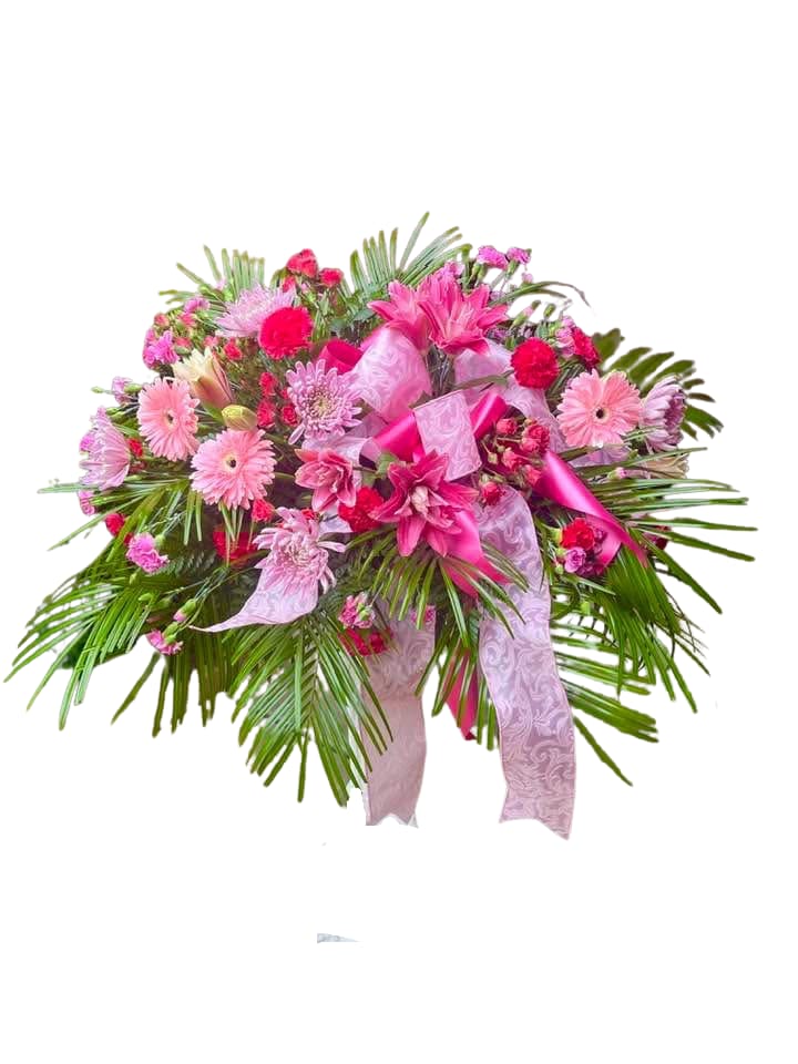 Rosado - A full, wide spray that reads as a graceful statement of care-Rosado is a memorial-style flower arrangement designed for sympathy, tributes, or any occasion that calls for a dignified show of support. The composition centers on pink lilies and gerbera daisies so the person you're honoring feels seen, surrounded by soft and vivid pinks that convey warmth and compassion. The arrangement's radiating palm fronds give it a formal, framed silhouette appropriate for services or to stand beside a keepsake at home.  This display layers soft and hot pink lilies, pink gerbera daisies, carnations, and cushion chrysanthemums to create a rich mix of textures-velvety petals, bold daisy faces, and rounded mums-set against vibrant green foliage. A patterned large pink bow sits at the heart of the spray with satin ribbons cascading down, lending a ceremonial finish. The piece is constructed wide and structured so it reads clearly from a distance and complements traditional memorial settings.  Choose Rosado when you want the recipient to feel comforted and honored; it's the kind of arrangement that communicates sympathy and steady support without words. Because it's styled as a spray with palms and long ribbons, please allow space for display and note that occasional trimming of stem ends and fresh water for the foam (if applicable) will keep blooms looking their best through services and viewings.