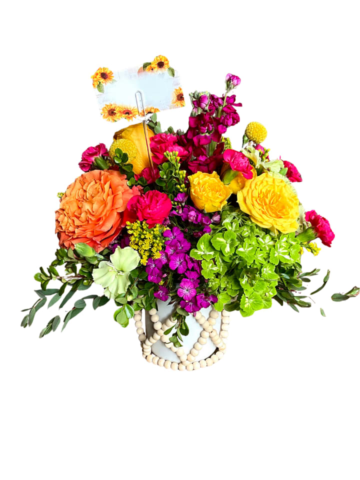 Bright Sentiments - Bold yellow and orange roses take center stage in this birthday bouquet, surrounded by vivid fuchsia and magenta blooms that create an immediate burst of color-perfect for birthdays, congratulations, or any moment you want to celebrate someone special. The arrangement reads joyful and energetic, the kind of gift that makes the person you're thinking of feel seen, celebrated, and a little giddy.  A dense cluster of roses, bright green hydrangeas, dianthus and stock mingle with small purple filler blooms and yellow accents for a lively, textured look. The flowers sit in a white ceramic vase wrapped in a geometric band of natural wooden beads, giving the piece a modern, handcrafted vibe. A decorative lemon and a floral-printed card on a metal pick add playful details that make the gift feel finished and festive.  This arrangement is ideal for sending to friends, family, or coworkers who deserve a cheerful lift-whether it's a milestone birthday, a new job, or simply "thinking of you." Standard care: keep in fresh water, away from direct heat and drafts, and refresh the water every other day to extend vase life.