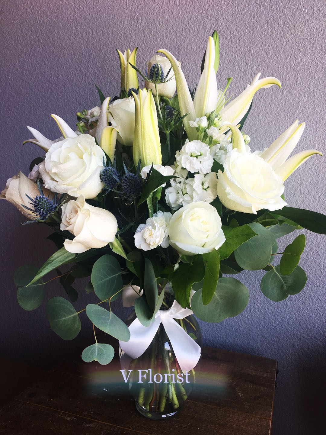 Tall arrangement of white lilies and roses with small white blooms in a clear glass vase with a white bow