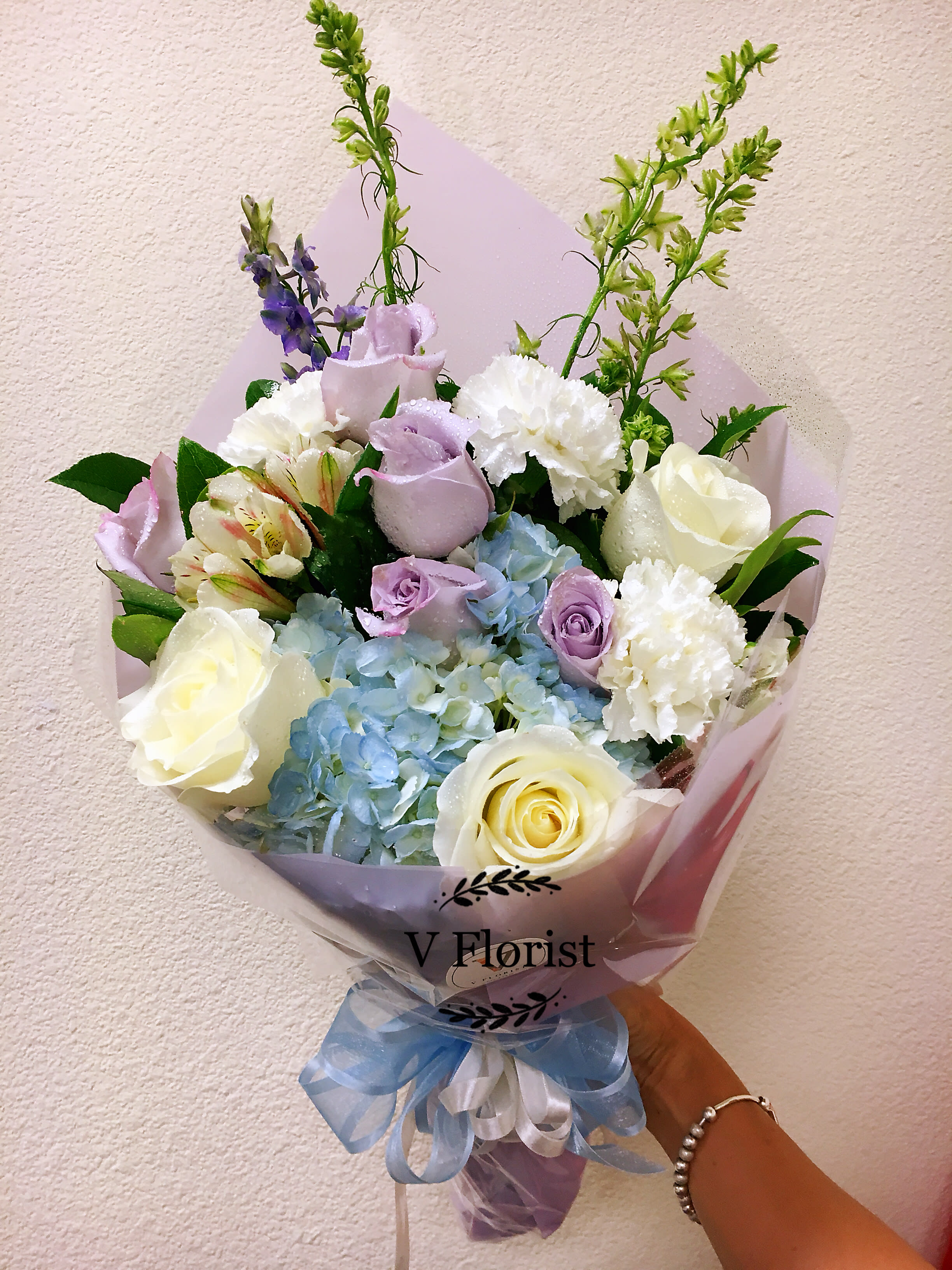 Hand-tied bouquet of white and lavender roses, blue hydrangeas, and white carnations wrapped in pastel paper.