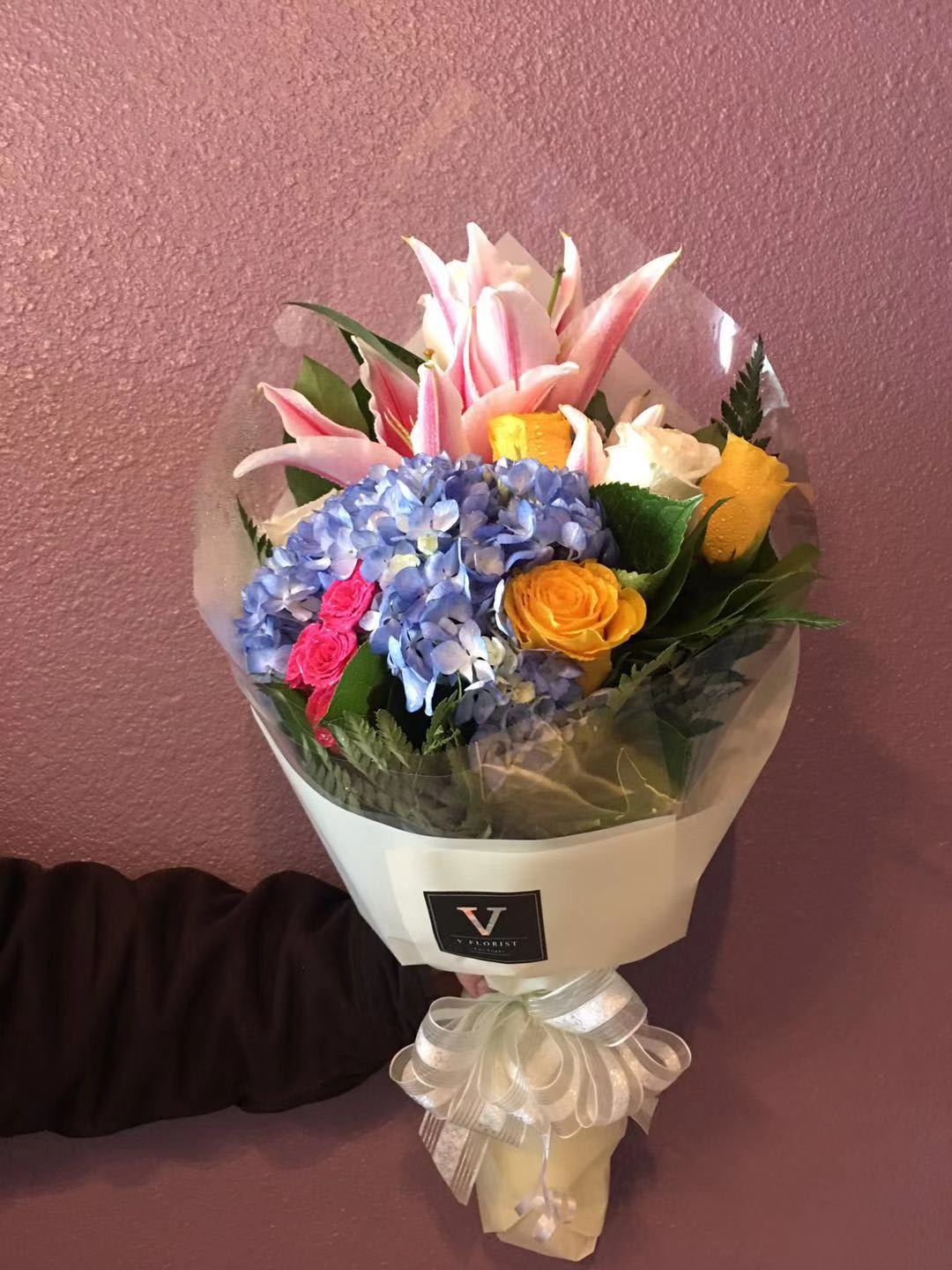 Hand-tied bouquet of pink lilies, blue hydrangea, and yellow and white roses in cream wrap with a bow.