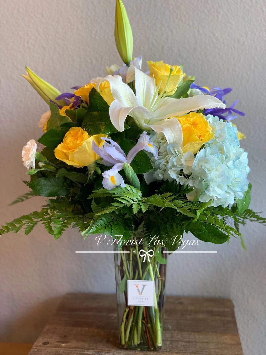 Tall arrangement of white lilies, yellow roses, blue hydrangeas, and purple iris in a clear glass vase