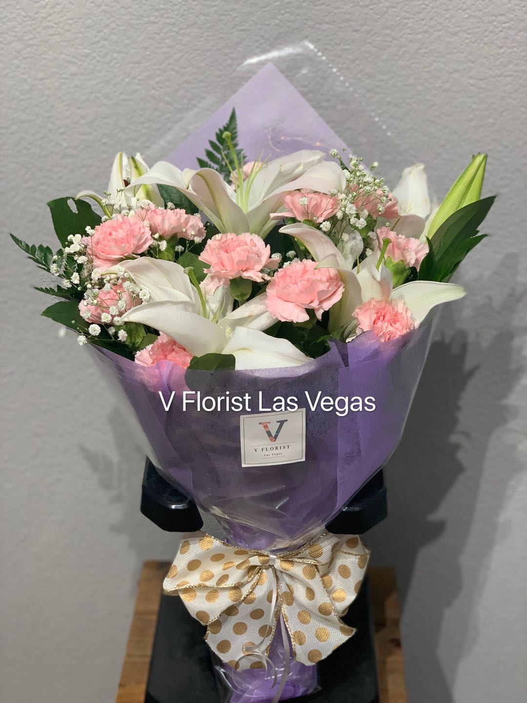 Hand-tied bouquet of white lilies and pink carnations wrapped in purple paper with a polka dot bow.