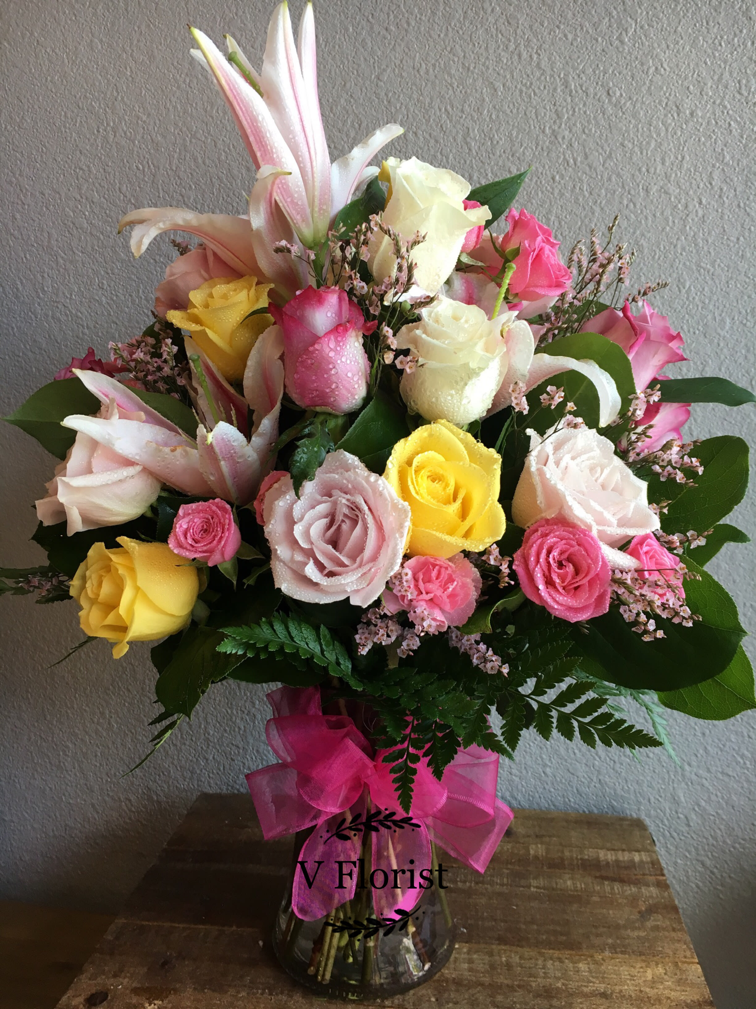Tall mixed arrangement of pink lilies and pink, yellow, and white roses in a clear vase with pink bow
