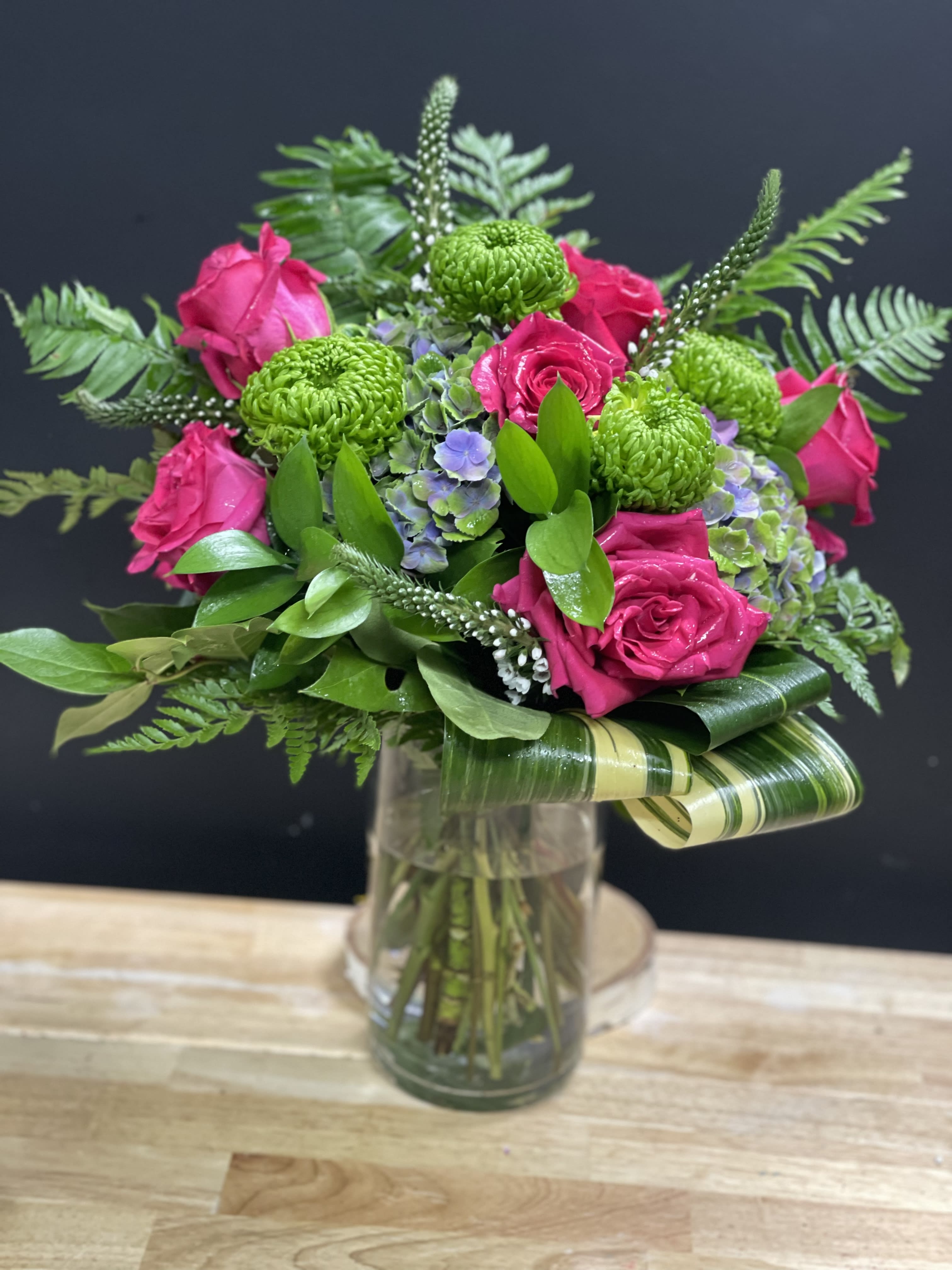 Vase arrangement of hot pink roses, green mums, and hydrangea with looped variegated leaves in a clear glass vase