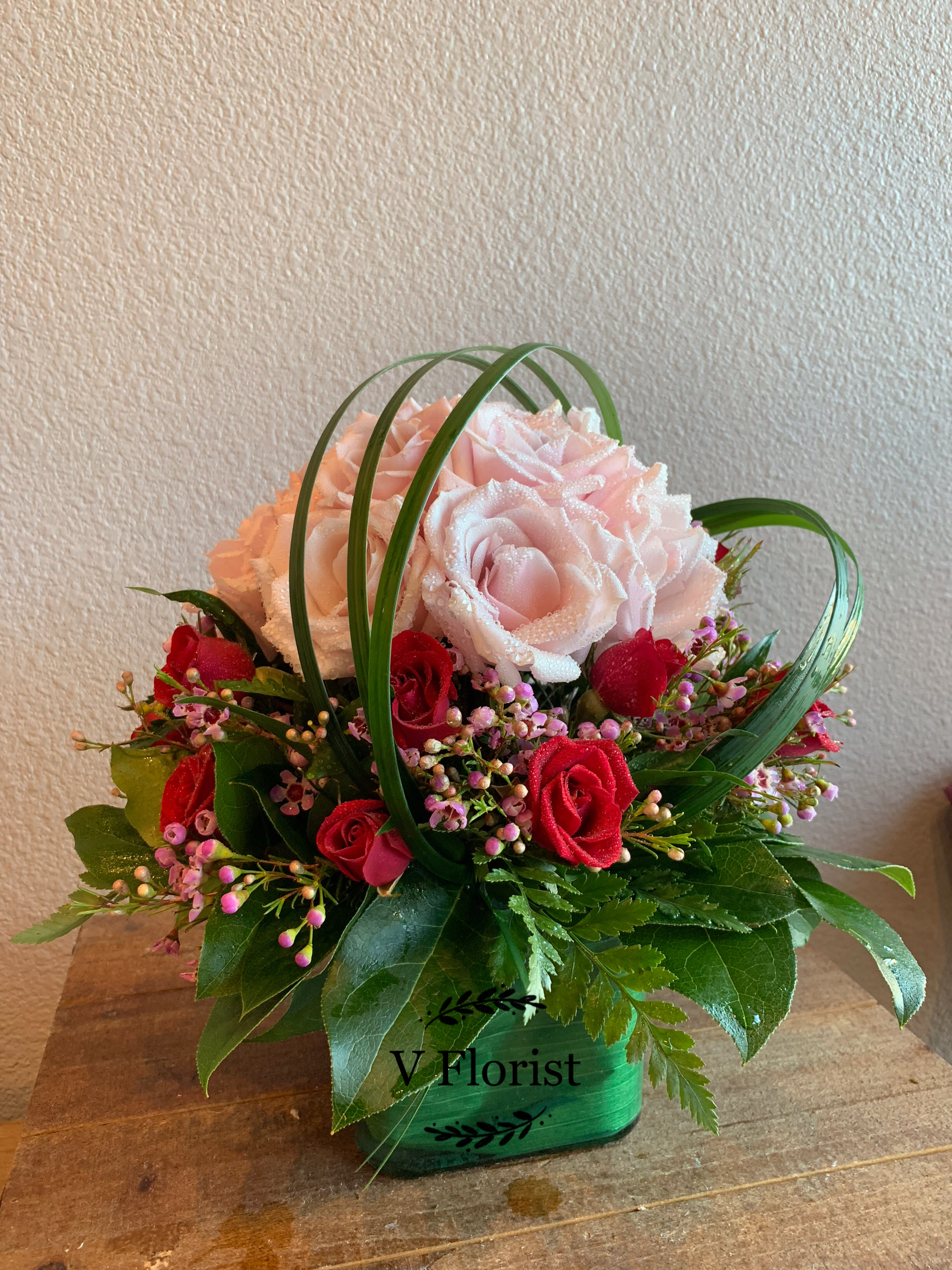 Short arrangement of pink and red roses with looping grass in a green cube vase