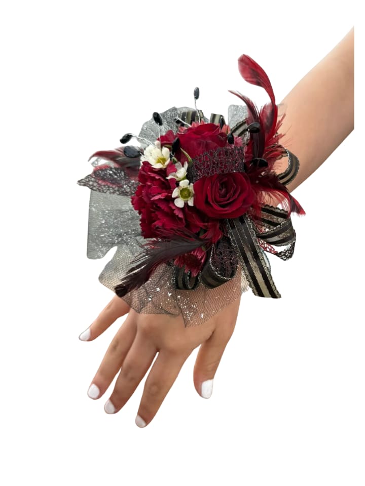 Rose and feather wrist corsage - Deep red roses and ruffled red carnations form a dramatic focal point in this wrist corsage, designed as an evening-ready accessory for proms, formal dances, or anniversary celebrations. The combination of red, black, and white - with small white daisy-like blossoms softening the palette - gives the recipient a bold, fashion-forward statement that reads both elegant and modern.  The flowers are arranged on a layered silver glitter mesh base that adds sparkle and volume around the wrist, tied with a black-striped ribbon for a tailored finish. Glossy black bead picks and sculptural red feathers introduce striking contrast and movement, making the corsage visible from every angle. Secured to a comfortable wrist band, it's sized to be worn with formalwear and coordinate with tuxes, evening gowns, or cocktail attire.  This is the kind of accessory that makes someone feel noticed and celebrated-perfect for the person you want to honor on a special night. Care: keep it cool and mist lightly if needed; order for same-day or event-date delivery to ensure peak freshness.