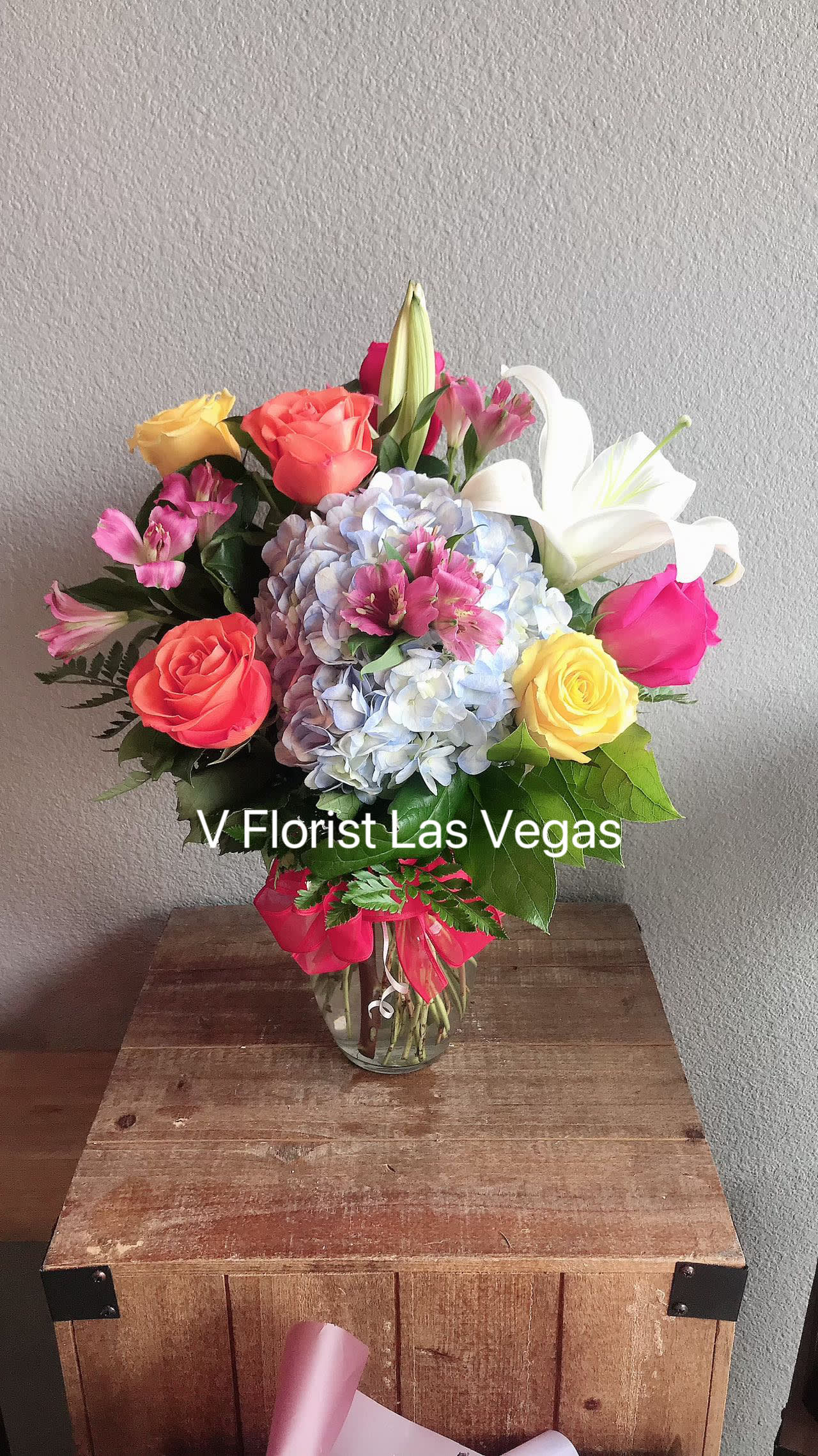 Bright mixed bouquet of roses, lilies, hydrangea and alstroemeria in a clear glass vase on a wooden crate.