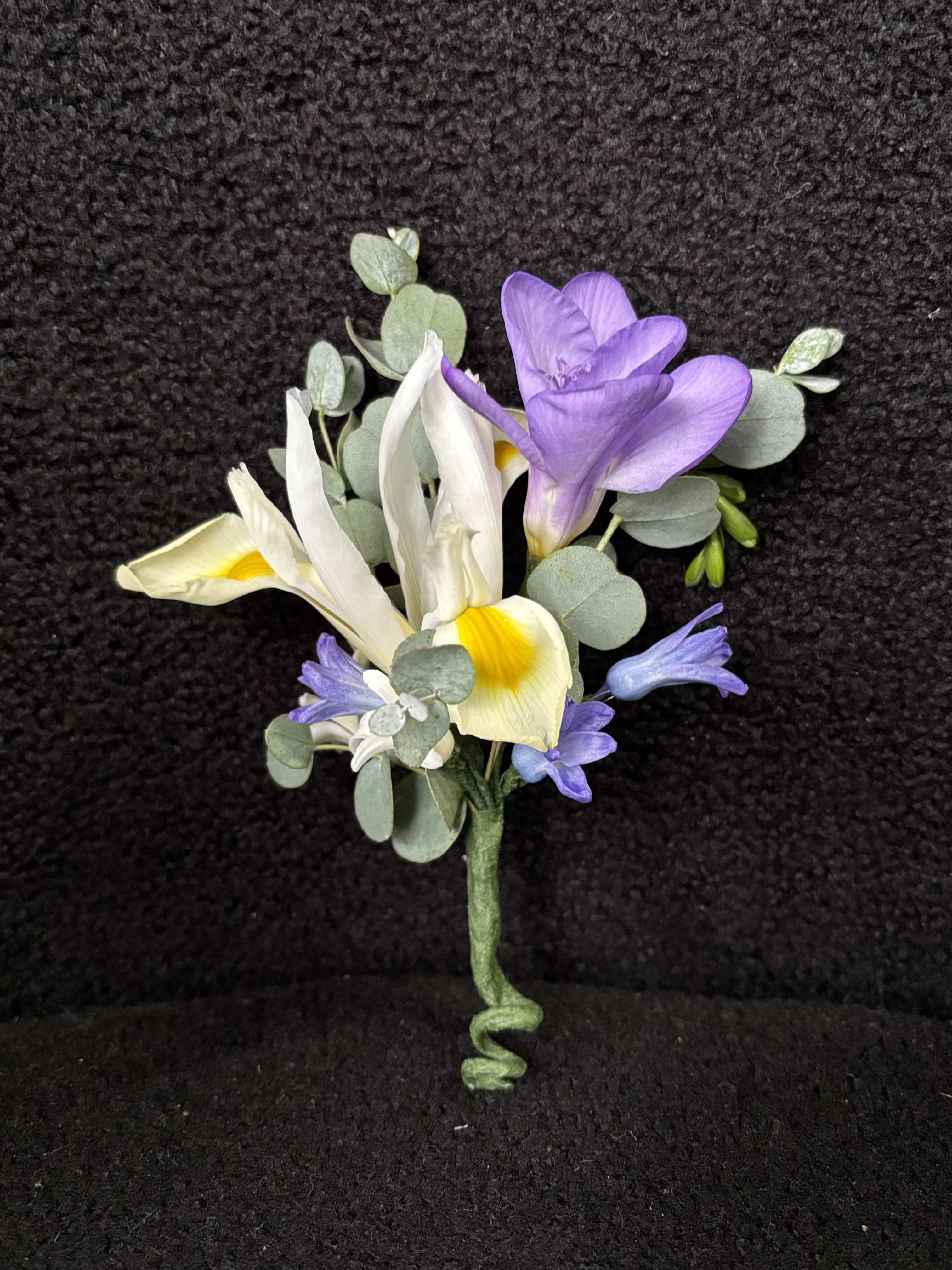 Whispering Garden Boutonniere - Whispering Garden Boutonnière is a refined floral accent designed to complement a romantic and elegant wedding aesthetic. Featuring delicate freesia, soft iris tones, and natural eucalyptus, this boutonnière brings a fresh, garden-inspired feel to the groom’s look. Light, airy, and thoughtfully composed, it enhances the overall style without overpowering. Each piece is carefully crafted to coordinate seamlessly with the bridal bouquet, creating a cohesive and sophisticated floral story. Finished with a clean stem wrap, it offers a timeless and polished presentation.
