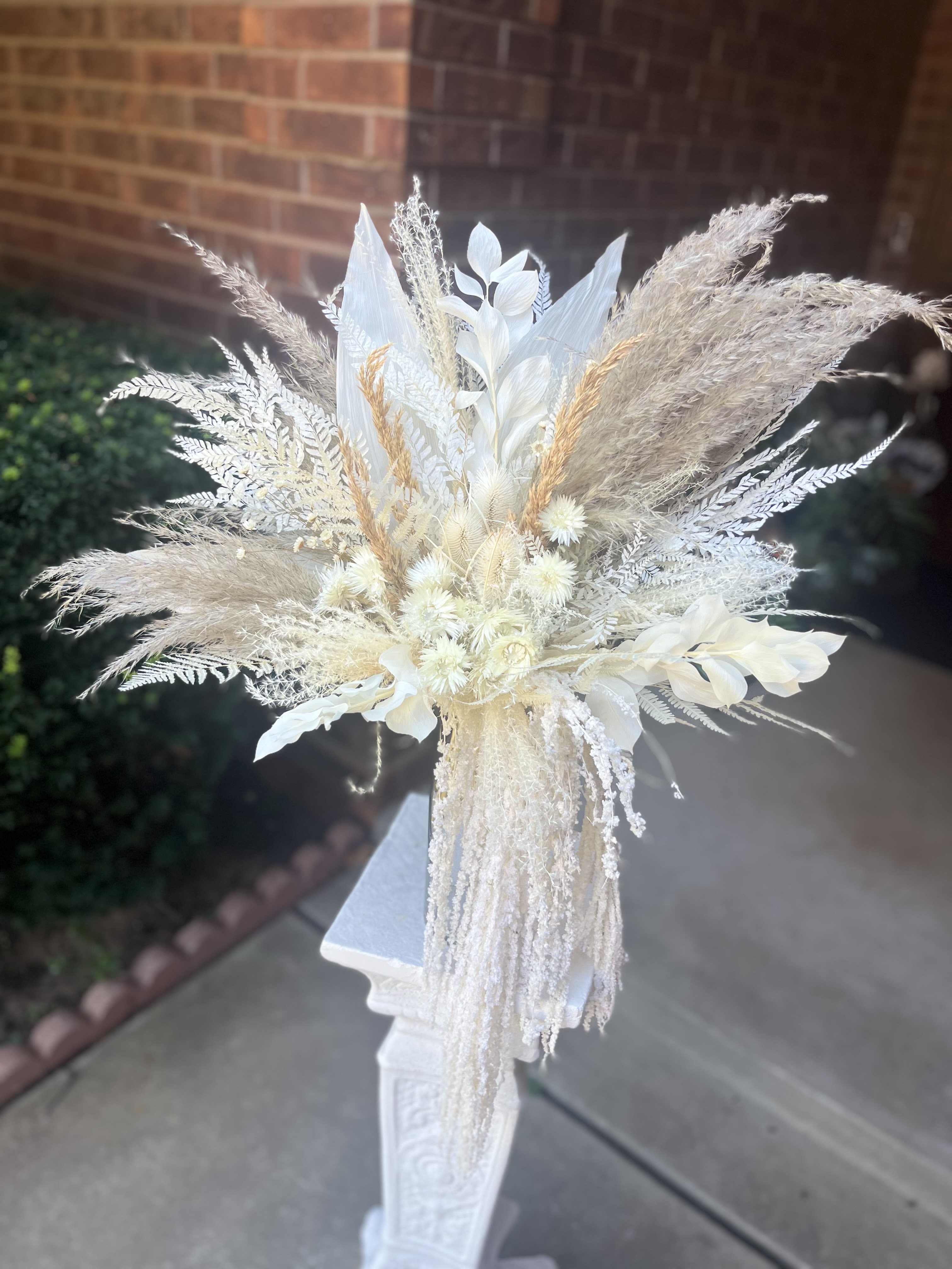 Dried Naturals Bouquet - An airy, sculptural statement for weddings: this dried naturals bouquet brings soft ivory, cream, and beige tones together in a modern, formal arrangement perfect for a bride seeking understated elegance or for ceremony and reception florals. As a wedding bouquet or altar piece, it reads timeless and refined-she'll feel centered and celebrated by its calm, architectural presence.  The bouquet features a mix of dried and preserved botanicals-tall pampas-style plumes that fan outward, bleached leaves and delicate dried ferns for structure, and cascading strands of fine textured material that create a flowing silhouette. Displayed here on an ornate white decorative pedestal, the composition emphasizes height and movement while keeping the palette neutral with whispers of white, yellow, and peach tones.  Ideal for minimalist, boho, or vintage-modern weddings and well suited to formal events or sympathy settings, this arrangement requires no water and keeps its shape season after season. Choose this piece when you want a lasting floral moment that looks intentional in photos and effortless in person; sizing and pedestal presentation make it especially effective as a ceremony focal or a sculptural reception accent.