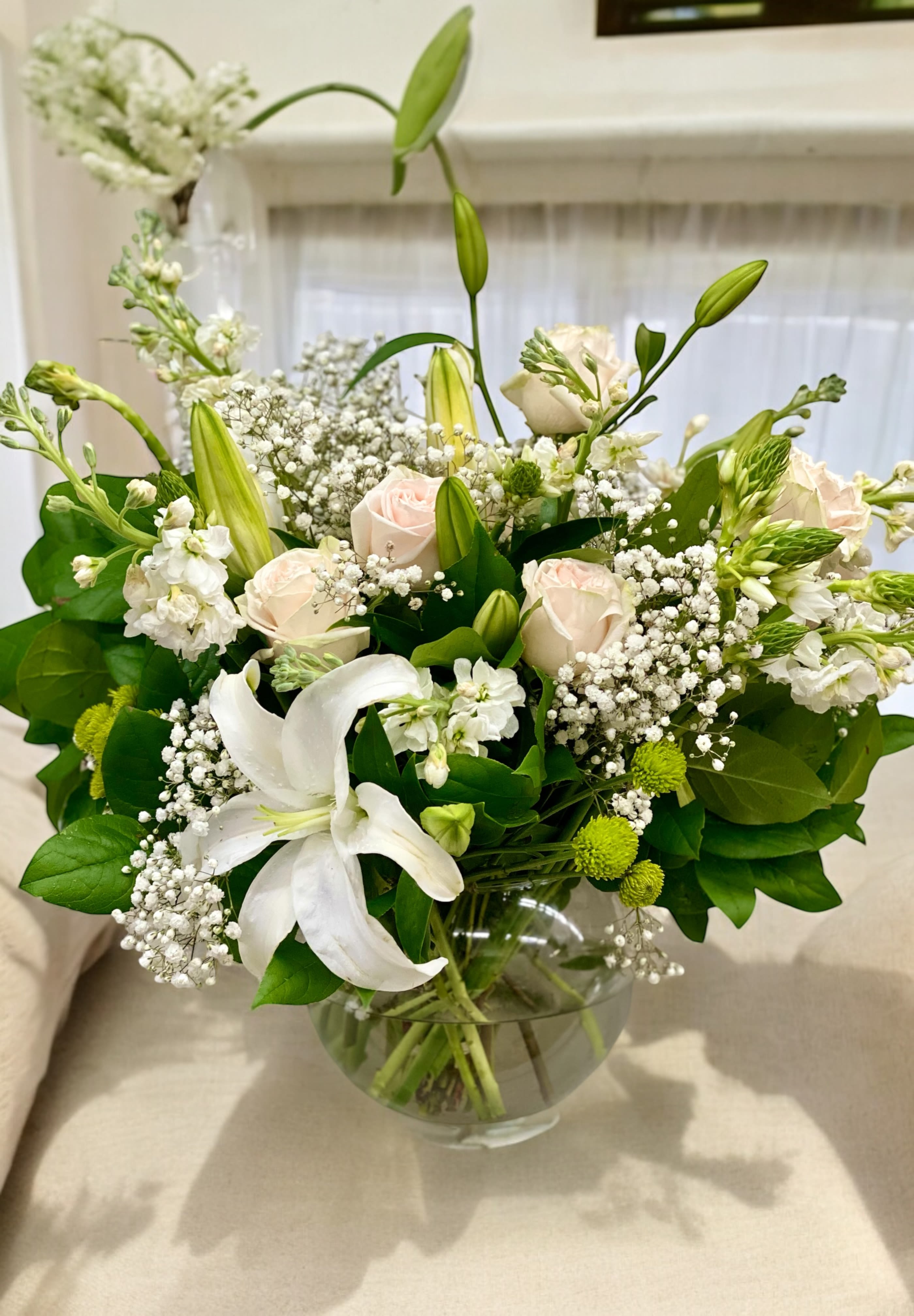 Sympathy White - A mixed arrangement of white lilies, blush roses, green button, white filler arranged in a clear round glass bowl.