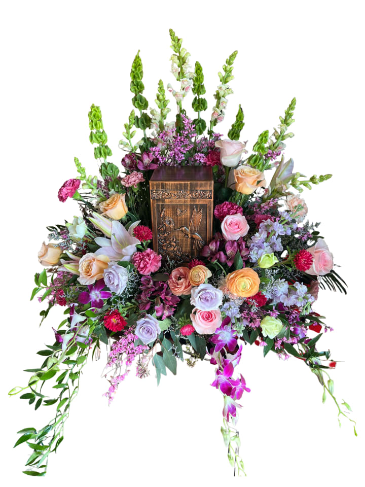 Deborah’s Garden - Soft pastels meet jewel-toned accents in this cremation flower arrangement crafted to comfort and honor - a memorial or celebration-of-life piece that puts a carved wooden urn at its heart. Deborah's Garden frames the urn in a lush surround so the person you're remembering feels cherished: delicate roses and ranunculus offer tender warmth, while lilies and carnations bring classic grace appropriate for an intimate service.  The design combines pinks, purples, peaches and fresh greens: dense clusters of roses, carnations, ranunculus, chrysanthemums and alstroemeria at the base; tall Bells of Ireland rising behind for gentle vertical presence; and cascading dendrobium orchids with trailing foliage to soften the composition. The carved wooden urn remains the focal point, supported by a richly textured mix of blooms that reads both elegant and reverent.  Perfect for a funeral home display or a at-home memorial, this arrangement conveys sympathy and thoughtful remembrance without overwhelming the space. The scale is intended to surround and honor a standard cremation urn; please contact us for delivery timing or to discuss personalization and care for delicate blooms during the service.  ***Urn not included